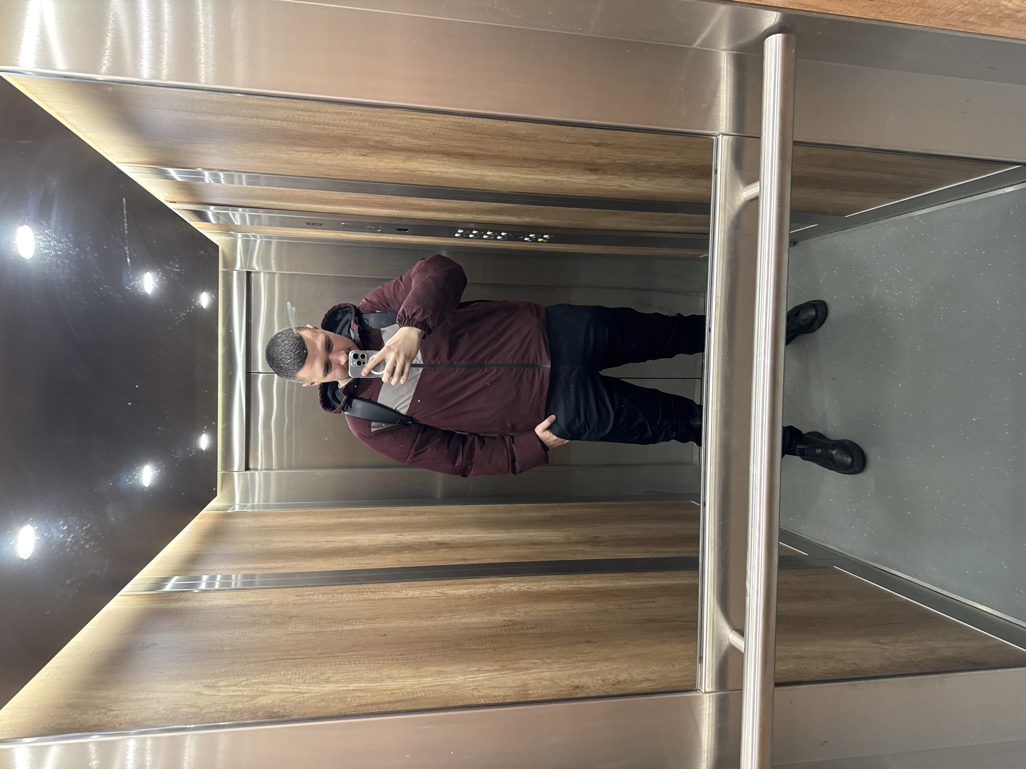 A man stands in the reflection of an elevator in Sofia, Bulgaria, taking a picture of himself with a smartphone. The elevator interior features wood paneling, stainless steel elements, and a dark panel with embedded lights. The man is wearing a maroon jacket with a grey stripe, black pants, and black shoes. The elevator floor is gray. Buttons are visible above the man's reflection in the elevator. A stainless steel handrail is visible to the right.