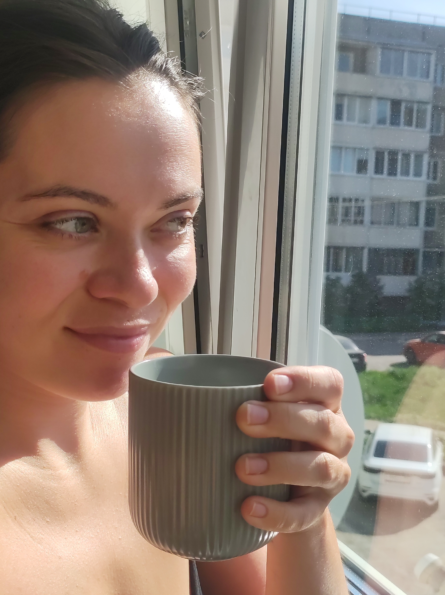 A woman stands by a window in Chaikovsky, Russia, holding a gray mug. She gazes out the window, a slight smile on her face. The background shows a multi-story apartment building, some parked cars, and greenery. The window appears to be a modern design with a white frame. The woman's hair is pulled back. The sunlight is bright.