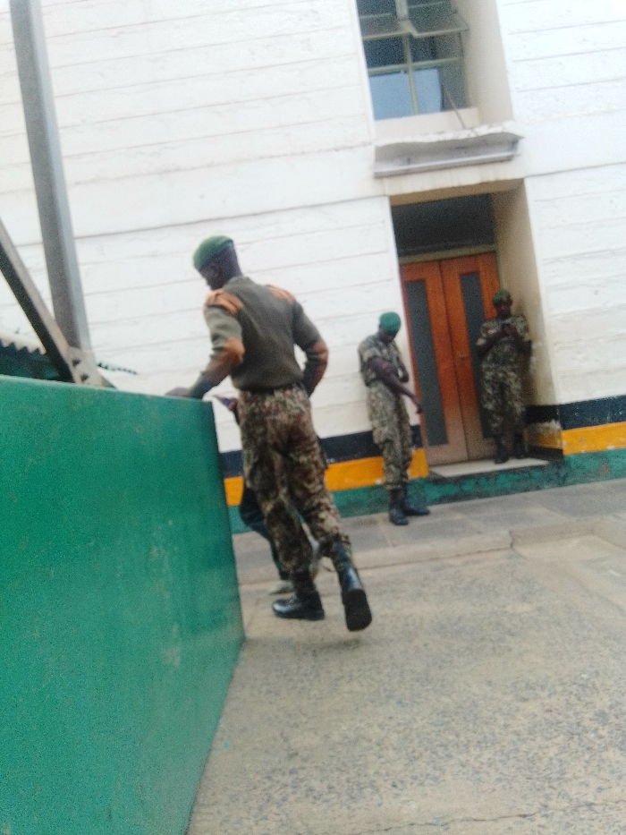 The image shows three individuals in military-style camouflage uniforms and green berets within what appears to be a secure compound or facility in Nairobi, Kenya. In the foreground, an individual with their back to the viewer wears an olive-green top with brown/gold shoulder epaulets, camouflage trousers, and black boots, with a green beret on their head. This individual is positioned leaning over a tall, matte-green barrier, with their left foot slightly raised as if in motion.
Behind this person, and further to the right, two additional individuals in similar camouflage uniforms and green berets stand near a light brown double wooden door, which is recessed into a light-colored building. The building wall features horizontal paneling or siding and an upper-level window. The base of the building's exterior wall is painted with prominent black, yellow, and green horizontal stripes. The ground surface is a grey, textured material, likely concrete. A metal support structure is partially visible behind the green barrier on the left side of the frame. One of the two background figures appears to be looking downwards, potentially at a handheld object. The scene suggests a controlled environment, consistent with a barracks or a government security installation.