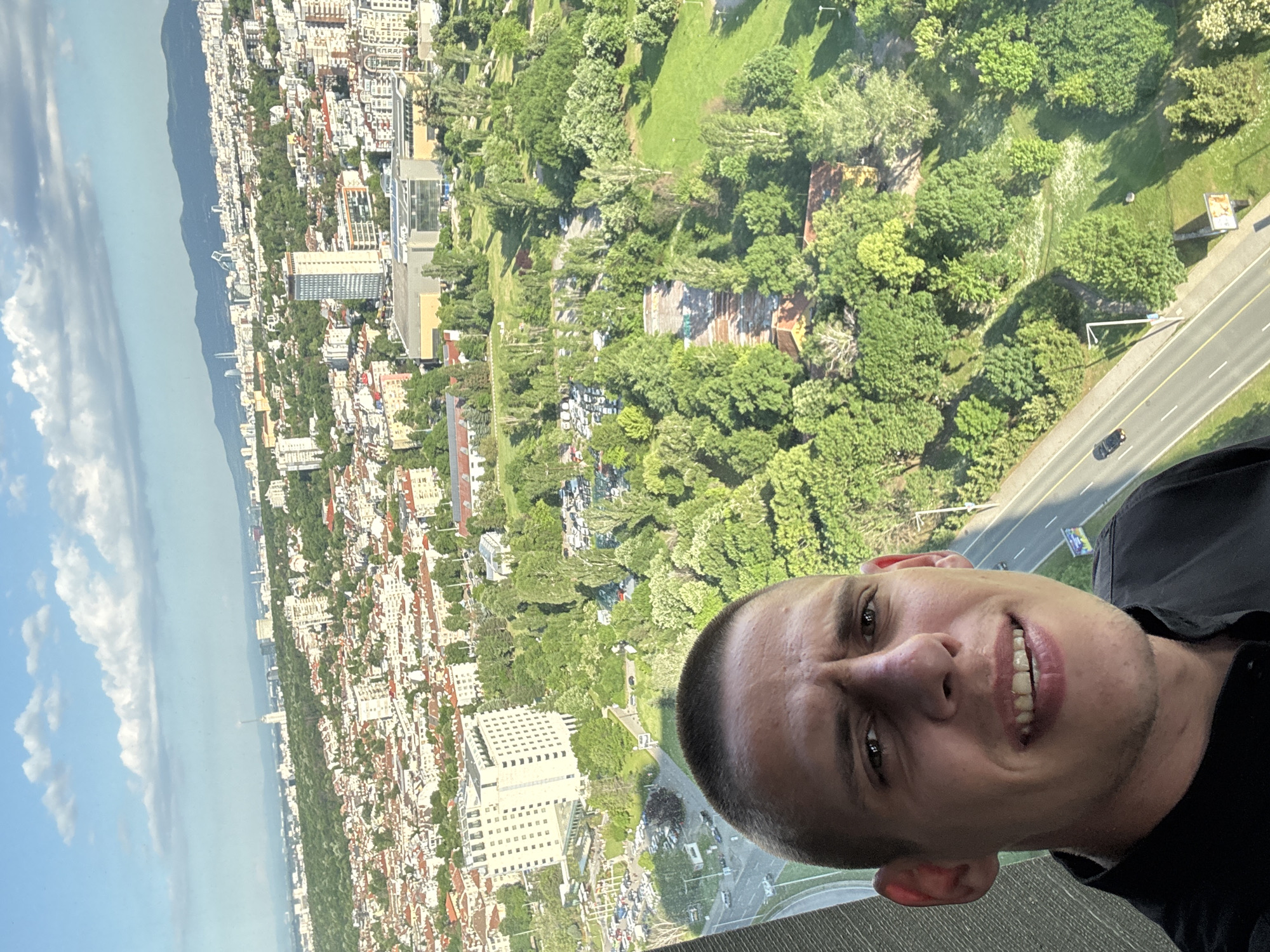 A photograph shows a man taking a selfie with a cityscape visible behind him. The scene outside the window appears to be the city of Sofia, Bulgaria. The view features residential and commercial buildings, trees, a highway, and a mountainous landscape in the distance. The sky is bright, with patches of clouds. The man is wearing a black shirt, and his expression is smiling.