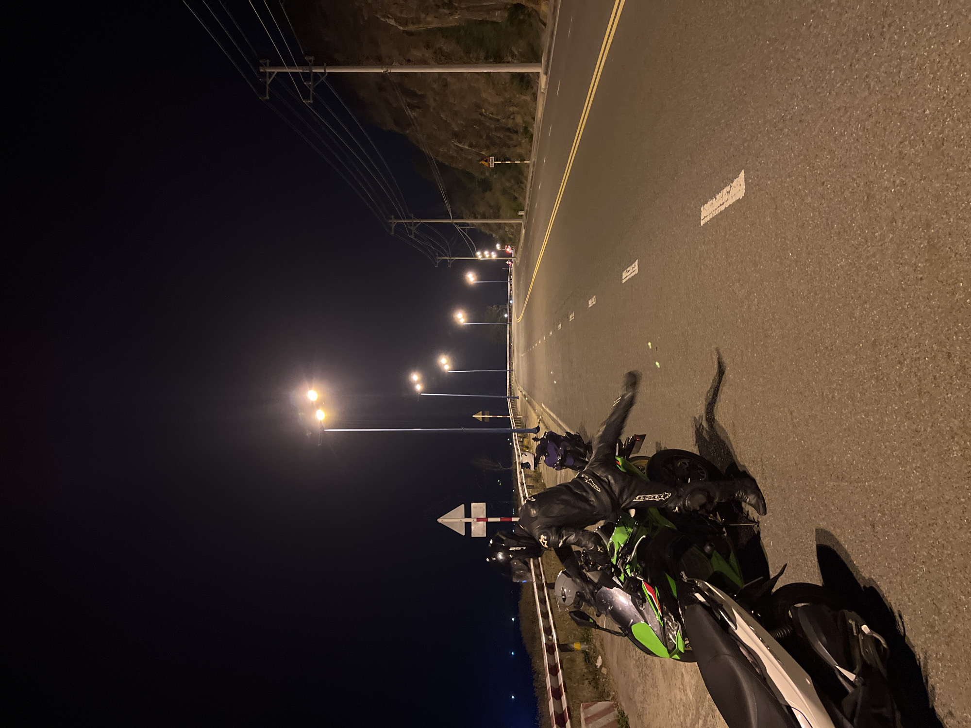 A motorcycle and its rider are on the shoulder of an asphalt road at night. The motorcycle is primarily green, black, and white. The rider is wearing a black leather suit and a helmet. The road has double yellow lines and intermittent white lines indicating lanes. Streetlights illuminate the road, and a series of utility poles line the left side. A hillside rises on the left. The scene is in the Bac Natural Heritage Area Trang, Vietnam.