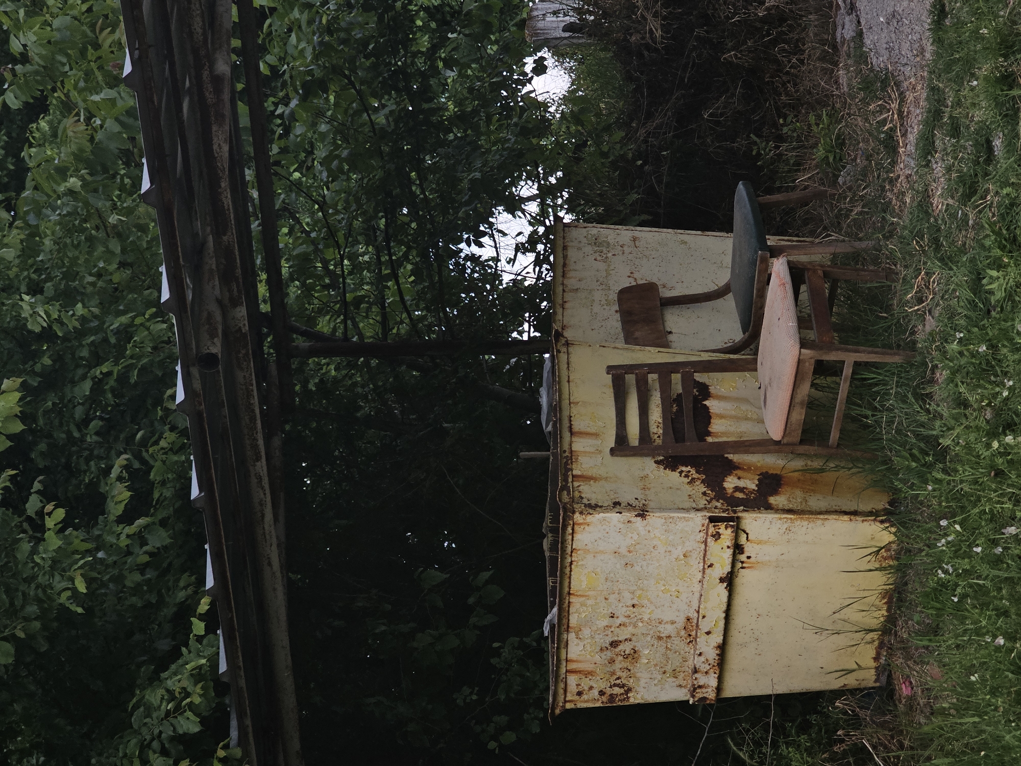 An outdoor scene in Dobri Dol, Bulgaria, shows two dilapidated metal boxes stacked on top of each other, heavily rusted and with peeling yellow paint. Two old wooden chairs, one with a green seat and the other with a pink seat, sit on top of the boxes. Lush green vegetation and grass surround the boxes on one side, while a metal structure stands on the opposite side, with dense trees and foliage filling the background.