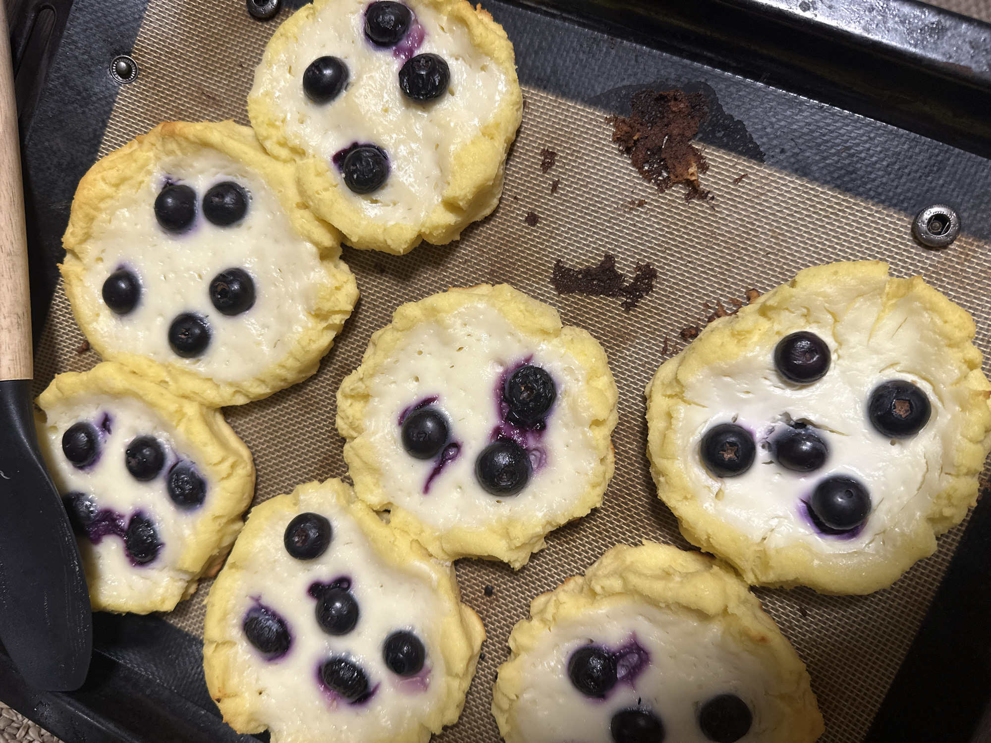 The image depicts a baking tray with seven blueberry and cream cheese pastries. The pastries are irregularly shaped with a pale yellow crust and a white filling dotted with blueberries, some of which have released purple juice into the filling. To the left of the pastries is a black spatula. A few crumbs of what appears to be burnt pastry are visible on the right side of the tray. The scene takes place indoors. There is no evidence to suggest that the image was taken in Vorotynsk, Russia.