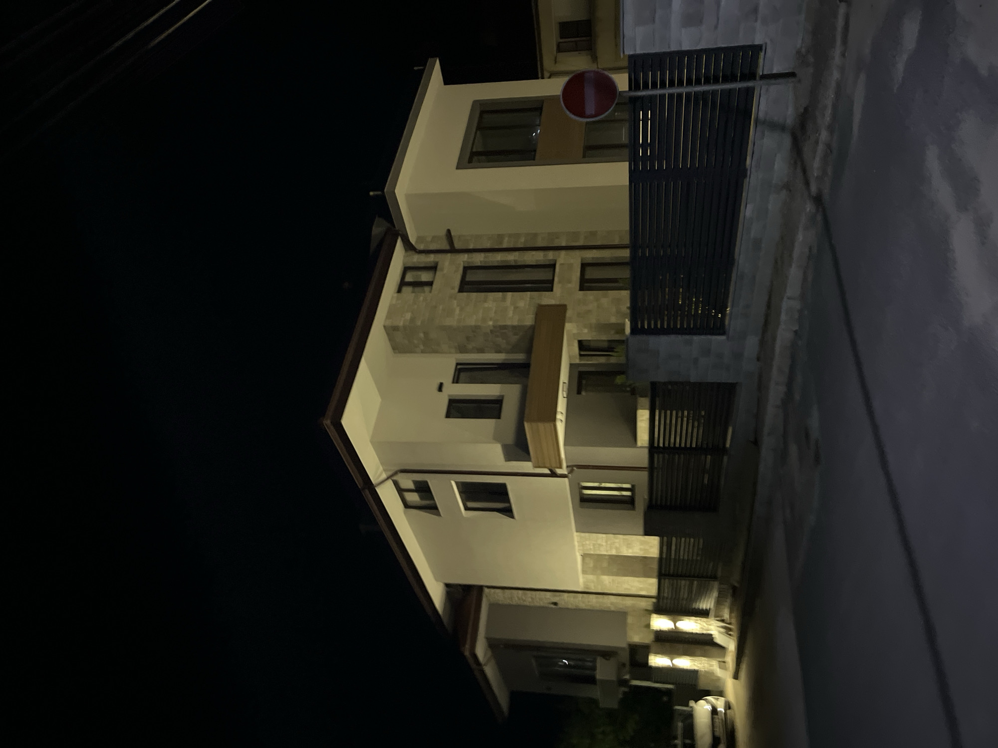 An exterior, eye-level, night-time, slightly low-angle shot shows a modern, multi-story house with grey and white stucco and stone accents, located on a street in Zlatograd, Bulgaria. The house features large windows, balconies, and a brown roof. A dark metal fence with horizontal slats encloses the property. A red and white circular "no entry" sign is visible on a metal pole to the right of the fence. To the left, a white car is parked near the entrance, illuminated by outdoor lights. The street in front of the house is asphalt and appears wet, reflecting the surrounding lights. Overhead, power lines are visible against the dark night sky.