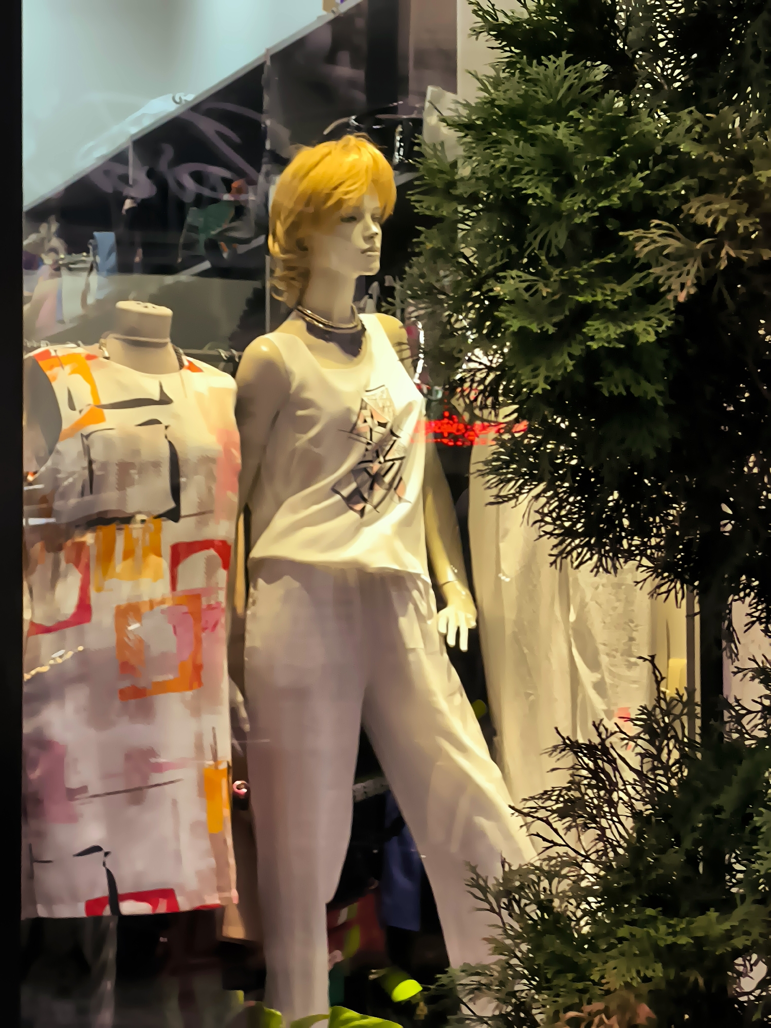 The image shows a store window display in Batumi, Georgia, featuring two mannequins dressed in casual attire. The mannequin on the left wears a sleeveless dress with a colorful abstract pattern of squares and rectangles in shades of pink, orange, yellow, and black against a white background. The mannequin on the right is dressed in a white sleeveless top with a black graphic design and light-colored pants. Both mannequins have blonde hair. A dark green coniferous shrub partially obscures the right side of the display. Reflections of the environment, including some red lights, are visible in the store window. The background appears to be the interior of a store with hanging garments.
