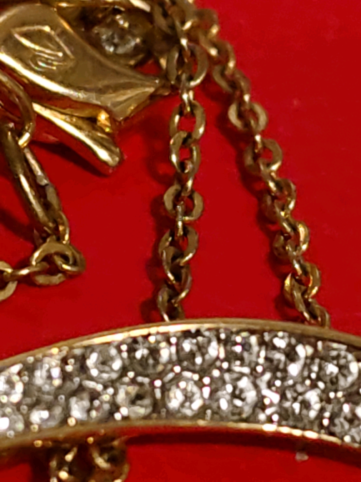 The image is a close-up of a piece of jewelry, likely a pendant or brooch, set against a bright red background. The jewelry features a gold-colored element with a detailed, possibly sculpted, design. This is connected to a delicate gold chain. A prominent part of the jewelry is an arc adorned with numerous small, clear stones, which appear to be rhinestones or diamonds, set in gold. The lighting creates reflections and highlights on the metallic and crystalline surfaces. There is no indication of the city of Rostov-on-Don, Russia, or any environmental context beyond the red surface.