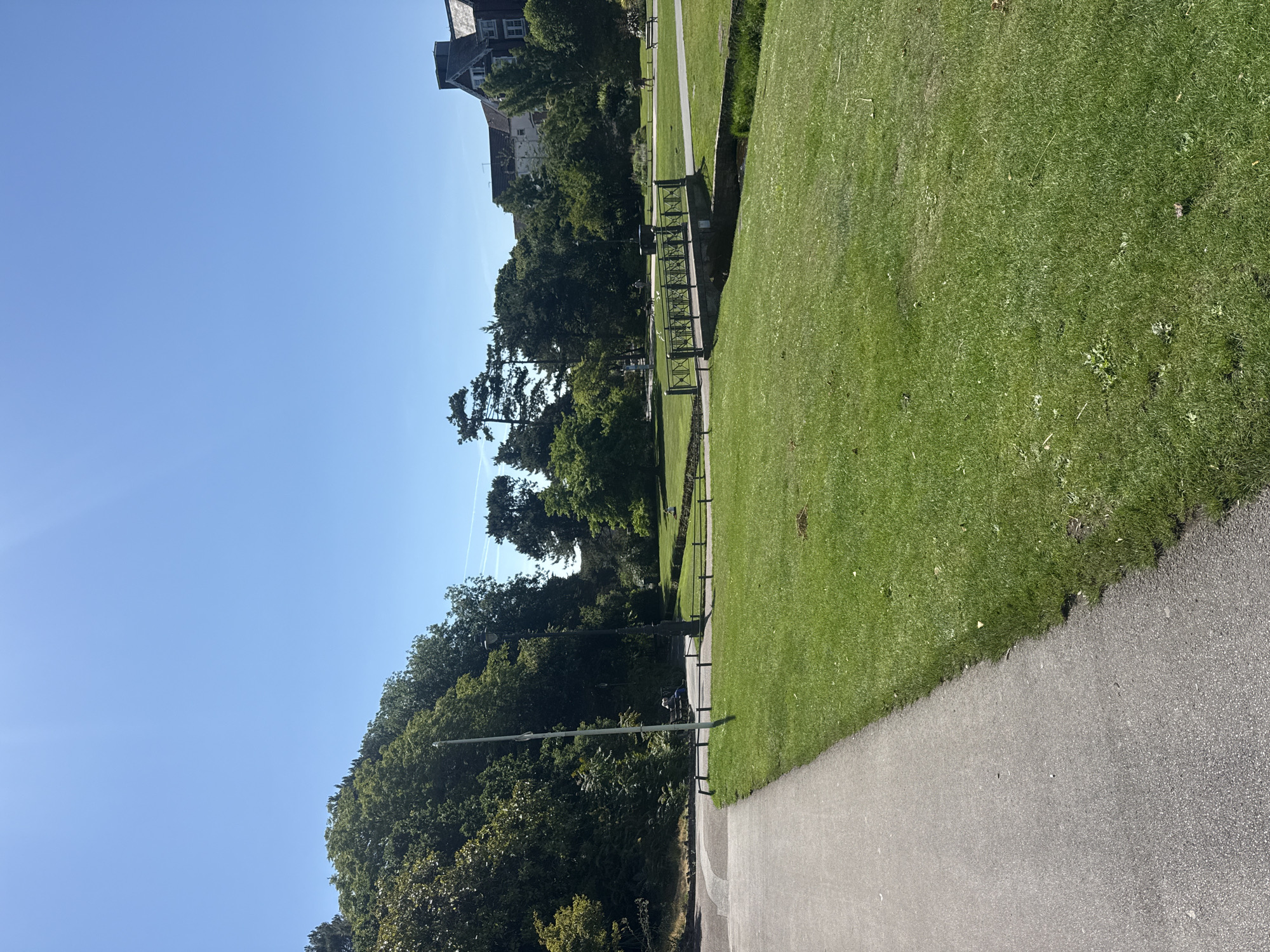 The image depicts a sunny day in a park, likely within Bournemouth, United Kingdom, given the architectural style of the buildings visible in the background. In the foreground, a paved pathway curves to the left, bordered by a well-maintained green lawn. Further into the park, a decorative metal bridge spans a narrow waterway. Lush trees and shrubs form a dense backdrop, with a few buildings peeking through the foliage. A lamppost stands near the pathway, and a bench is occupied by a person sitting with their back to the camera. Several other individuals are seen walking in the distance, enjoying the park. The sky is a clear, bright blue, suggesting fair weather.
