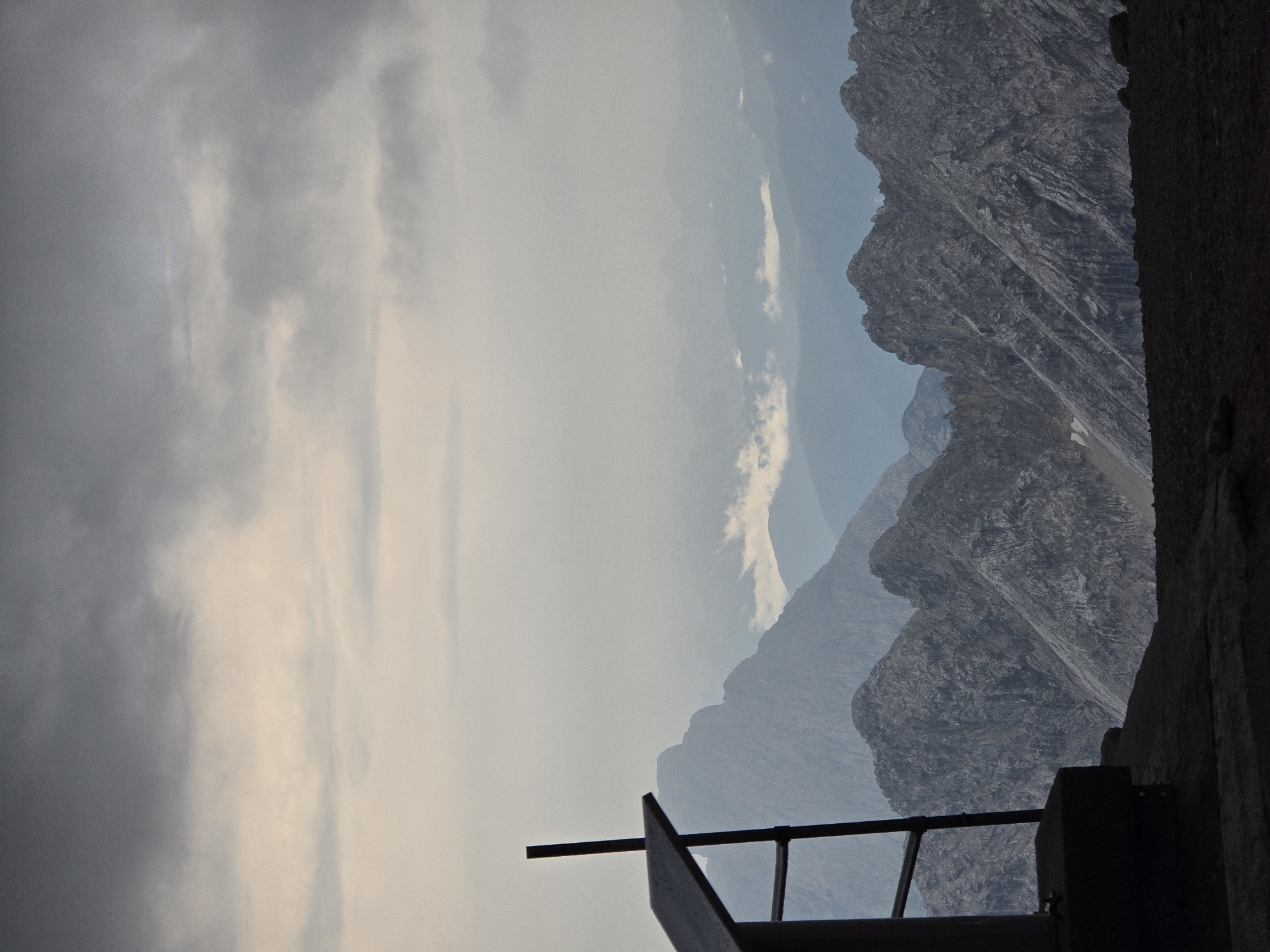 This image depicts a panoramic view of rugged, mountainous terrain under a cloudy sky. In the foreground, a dark, metallic structure with a vertical post and a slanted surface, possibly a railing or a sign, is positioned to the left. The structure is silhouetted against the distant landscape.
The middle ground is dominated by sharp, rocky mountain peaks, characterized by their grey, textured surfaces. The mountains recede into the distance, becoming increasingly blurred and appearing in shades of blue and grey due to atmospheric perspective. Wisps of clouds are visible nestled between some of the mountain ranges.
The sky above is overcast with dense, dark grey clouds, suggesting a somber or potentially stormy atmosphere. The lighting is diffused, creating a muted palette across the entire scene.
While the exact location is not visible in the image, this type of alpine landscape is characteristic of the area surrounding Garmisch-Partenkirchen, Germany, a region known for its dramatic mountain ranges, including the Zugspitze.