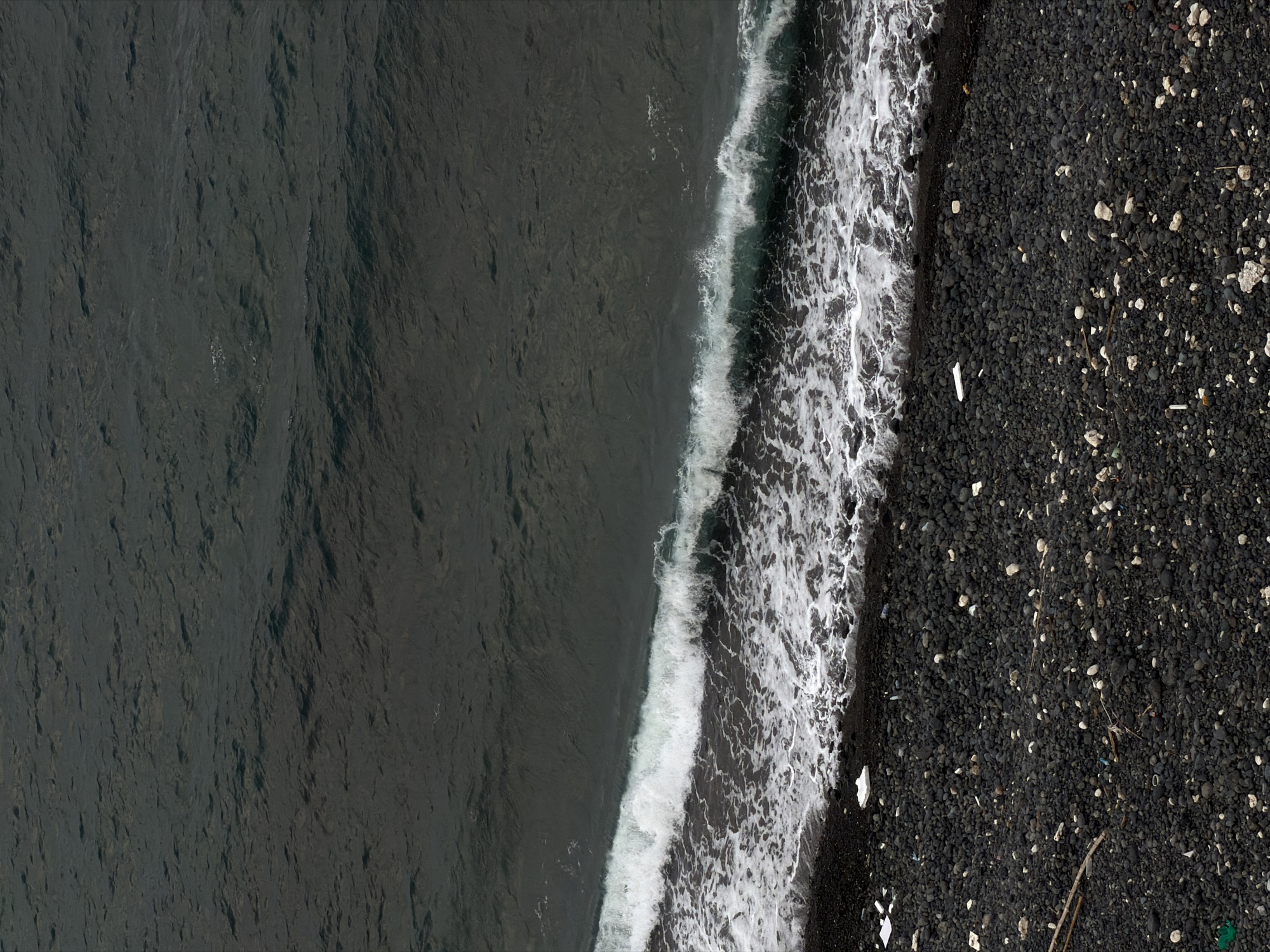 The image shows an aerial view of a black sand beach in Nam Nha Trang Ward, Vietnam. The left side of the image is the dark, rippled ocean, with a white foamy wave crashing along the shoreline. The right side is the black sand beach, sparsely scattered with small white and grey rocks, debris, and a few pieces of thin sticks or twigs. The perspective is directly overhead, looking down onto the beach and water.
