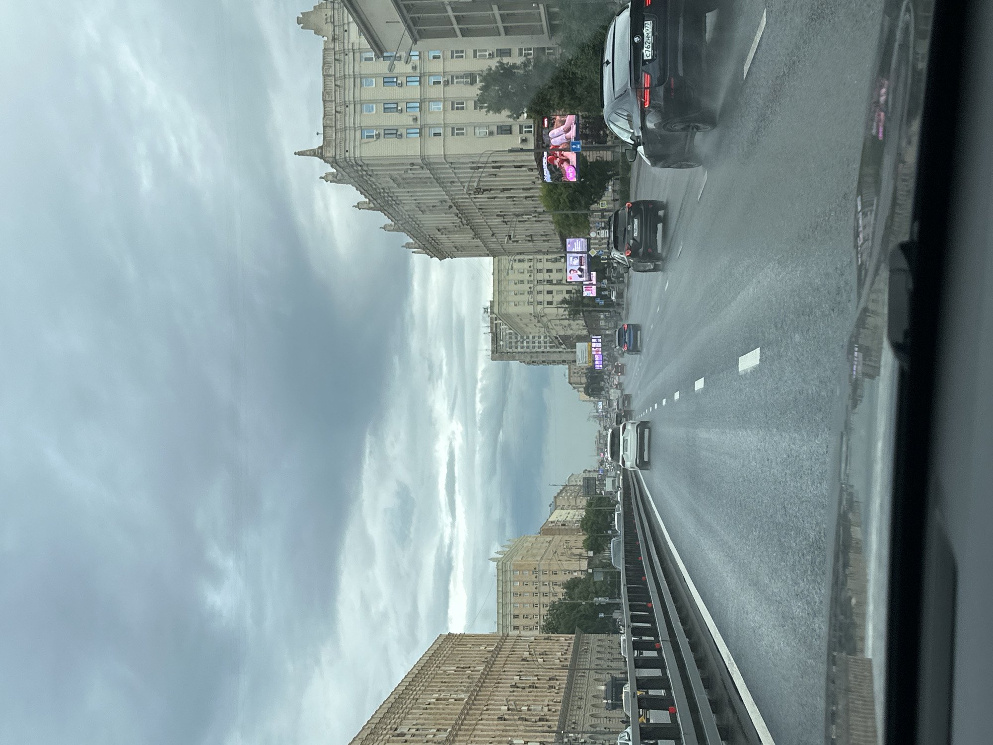 The image depicts a street scene in Moscow, Russia, captured from inside a vehicle. The perspective is tilted, suggesting the photo was taken from a car moving along a multi-lane road. The sky is overcast with dramatic, heavy clouds, indicating a cloudy or possibly rainy day.
On the left side of the road, large, imposing buildings with many windows line the street. These buildings appear to be residential or office complexes, showcasing distinct architectural styles typical of some Moscow urban areas, with ornate facades and classical elements. Some of these buildings are quite tall, reaching several stories high.
On the right side of the road, traffic is moving. Several cars are visible, including a black BMW SUV in the foreground with a license plate that appears to read "C762HM97". The road surface is wet, suggesting recent or ongoing precipitation. A white vehicle is visible further down the road, also in traffic. Digital billboards are mounted on poles along the street, displaying colorful advertisements, some of which appear to feature people.
The overall impression is that of a busy urban environment during daytime, under a gloomy sky. The wet road and overcast conditions contribute to a somewhat muted atmosphere. There are no discernible emotions conveyed, as the focus is on the urban landscape and traffic. The visible text on the license plate and billboards provides specific details about the location and potential advertisements.