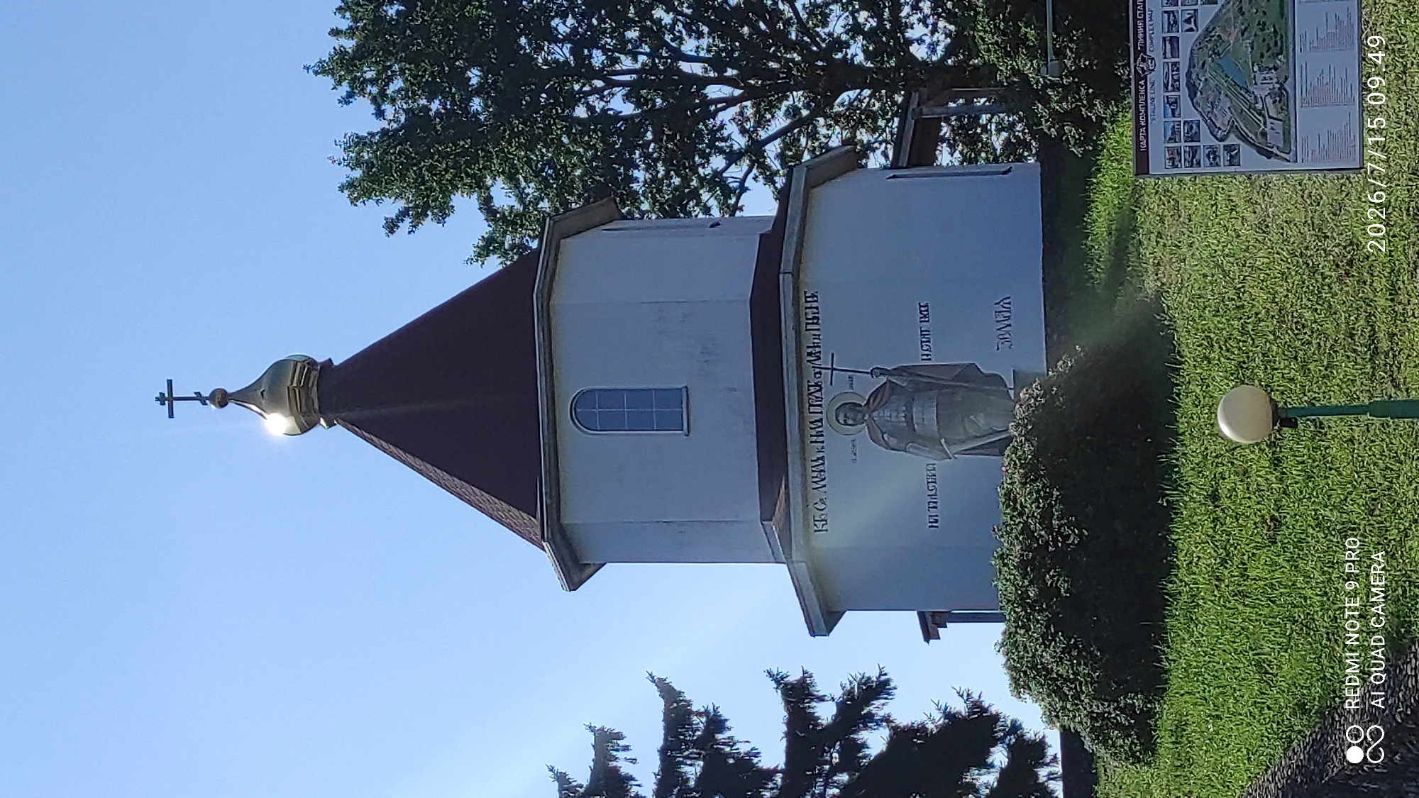 This is a medium-shot, outdoor, eye-level, vertical photograph of a white, octagonal church or chapel with a dark brown, conical roof topped by a golden dome and a cross. The building is situated on a grassy hill. A statue of a robed figure holding a cross is painted on the lower section of the building. There is text painted above and below the statue, some of which is in Cyrillic script. To the left of the building, a large evergreen tree with dark green needles is partially visible. To the right, the lush green canopy of a deciduous tree is seen against a clear blue sky. In the foreground, a portion of a grassy slope is visible, with a short, dark green bush in front of the statue. A white, spherical light fixture on a green pole stands in the lower right quadrant of the image. In the bottom right corner, a map is affixed to a post. The bottom of the image displays a timestamp "2026/7/15 09:49" and phone information "REDMI NOTE 9 PRO AI QUAD CAMERA". The scene appears to be located in or near the city of Лашанскі сельскі Савет, Belarus.