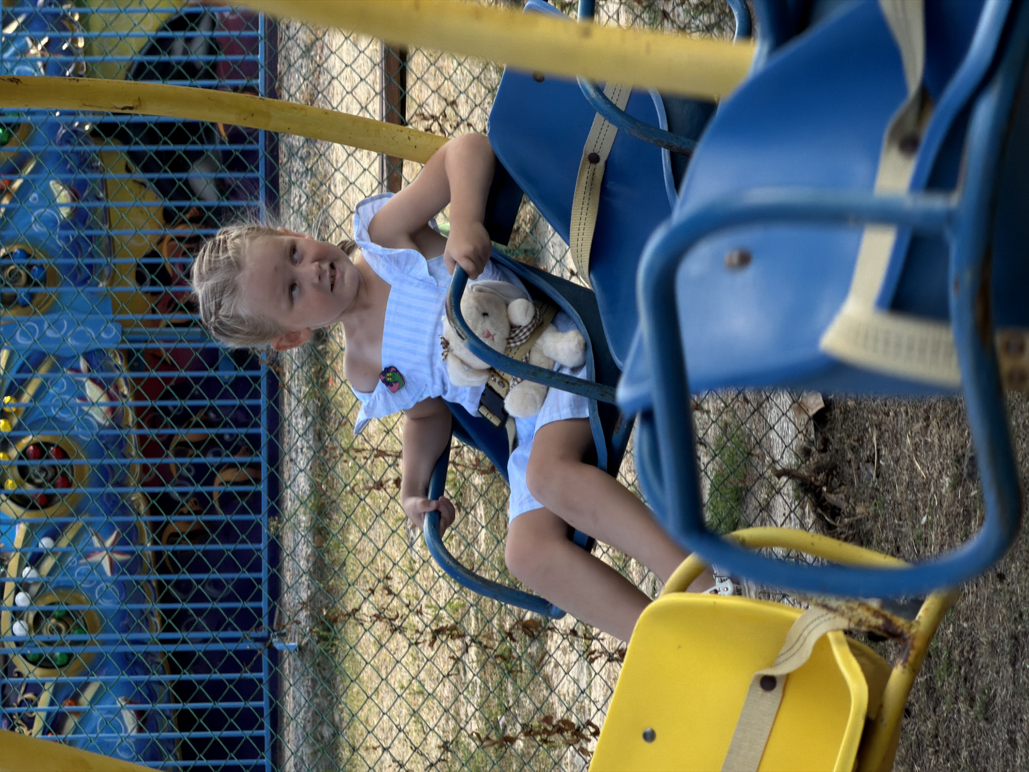 A young Caucasian girl with blonde hair braided into a ponytail sits on a blue swing seat. She is wearing a white and blue striped dress with ruffled sleeves. She holds a small, light-colored stuffed bunny with a checkered scarf around its neck. The girl is looking to her right with a slight smile. In front of her, another blue swing seat and a yellow swing seat are visible. The swings are part of a playground structure, with a blue metal fence in the background and dry grass and dirt on the ground. A colorful, blurry amusement park ride is visible behind the fence. The scene is outdoors, likely in a park or amusement area in the city of Kamyshin, Russia.