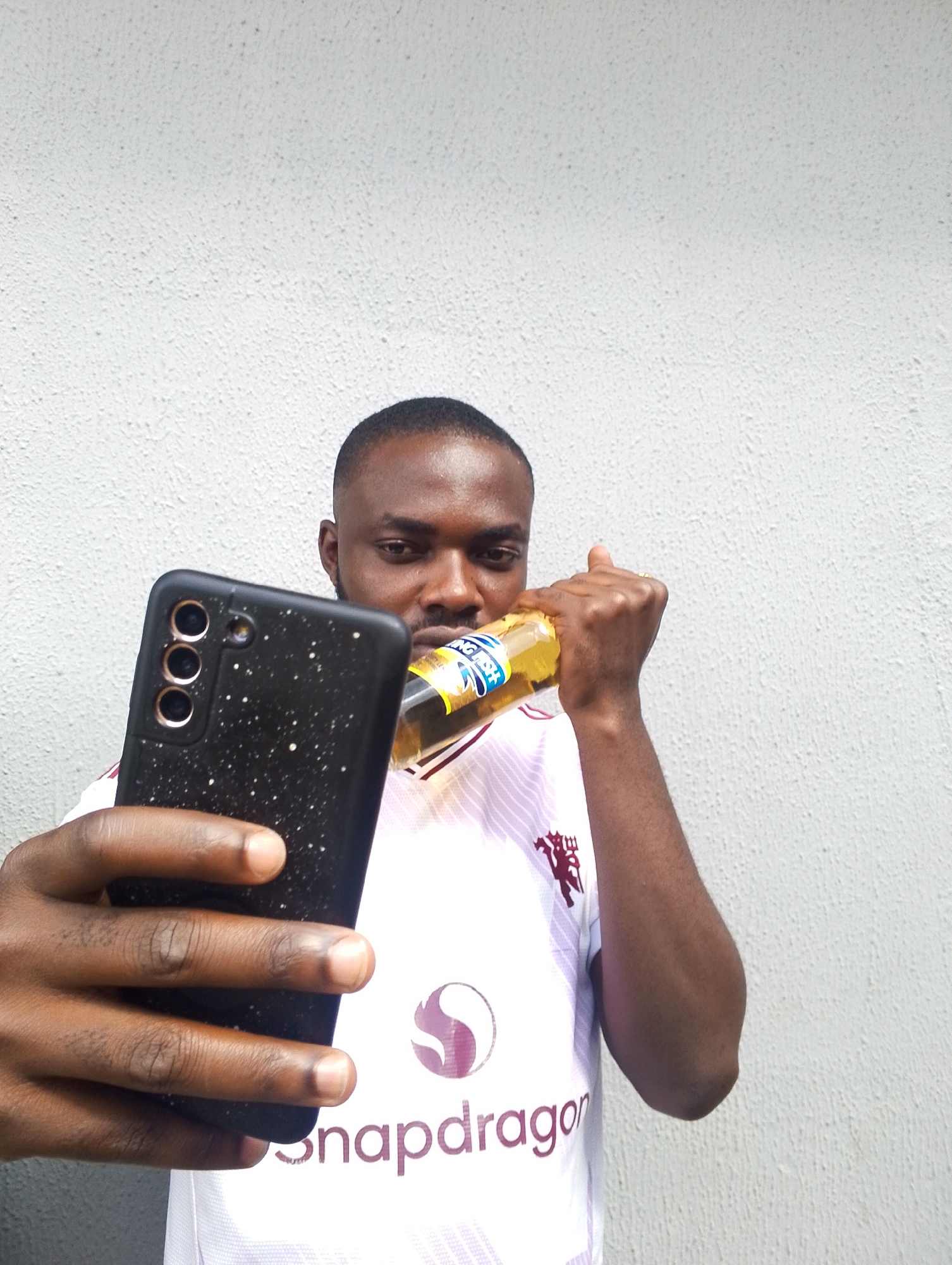 A young Black man is taking a selfie in front of a textured white wall. He is holding a black smartphone in his left hand, with his thumb resting on the screen. His right hand is holding a bottle of drink to his mouth, tilted slightly as if he is taking a sip. He is wearing a white jersey with maroon accents and logos, including a prominent "snapdragon" logo at the bottom. The jersey also features a crest on the upper right chest. The man has short-cropped hair and is looking directly at the camera. The lighting suggests it is daytime. The background is plain, indicating an outdoor setting or a room with a blank wall. There are no clear indications of the specific time of day or weather, but the lighting is even. The image is taken from the chest up, focusing on the man, his phone, and the drink.