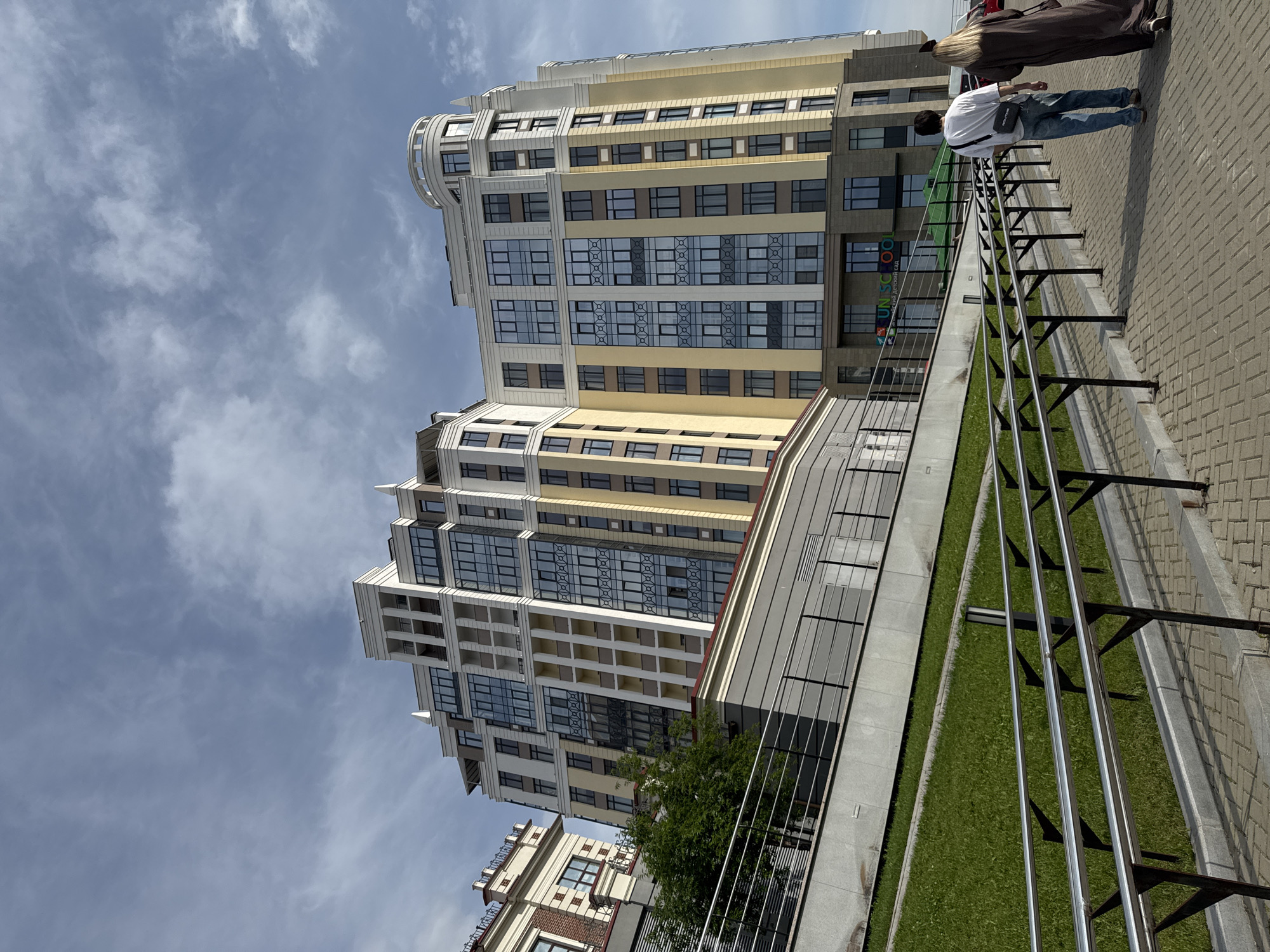 Two people walk on a paved pathway in front of a large, modern apartment building in Yekaterinburg, Russia. The building is tall and multi-storied, with a beige and yellow facade, and numerous windows. A grassy area slopes up to the left of the pathway, bordered by a metal railing. The sky is blue with scattered clouds. The building has "UNISCHOOL" signage above its entrance.
