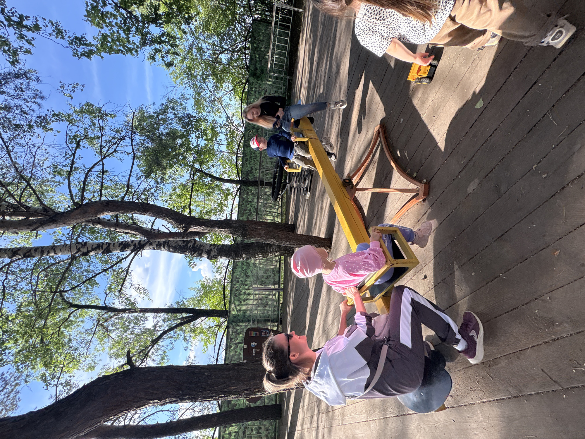 The image captures a scene at a playground in Chaikovsky, Russia. Four individuals are visible, with three of them on a yellow seesaw. A woman in athletic wear sits on one end of the seesaw, while a child in a pink outfit sits on the other end. Two other people sit on the other seesaw. The playground is situated on a wooden deck surrounded by trees. The sky is partially visible through the foliage.