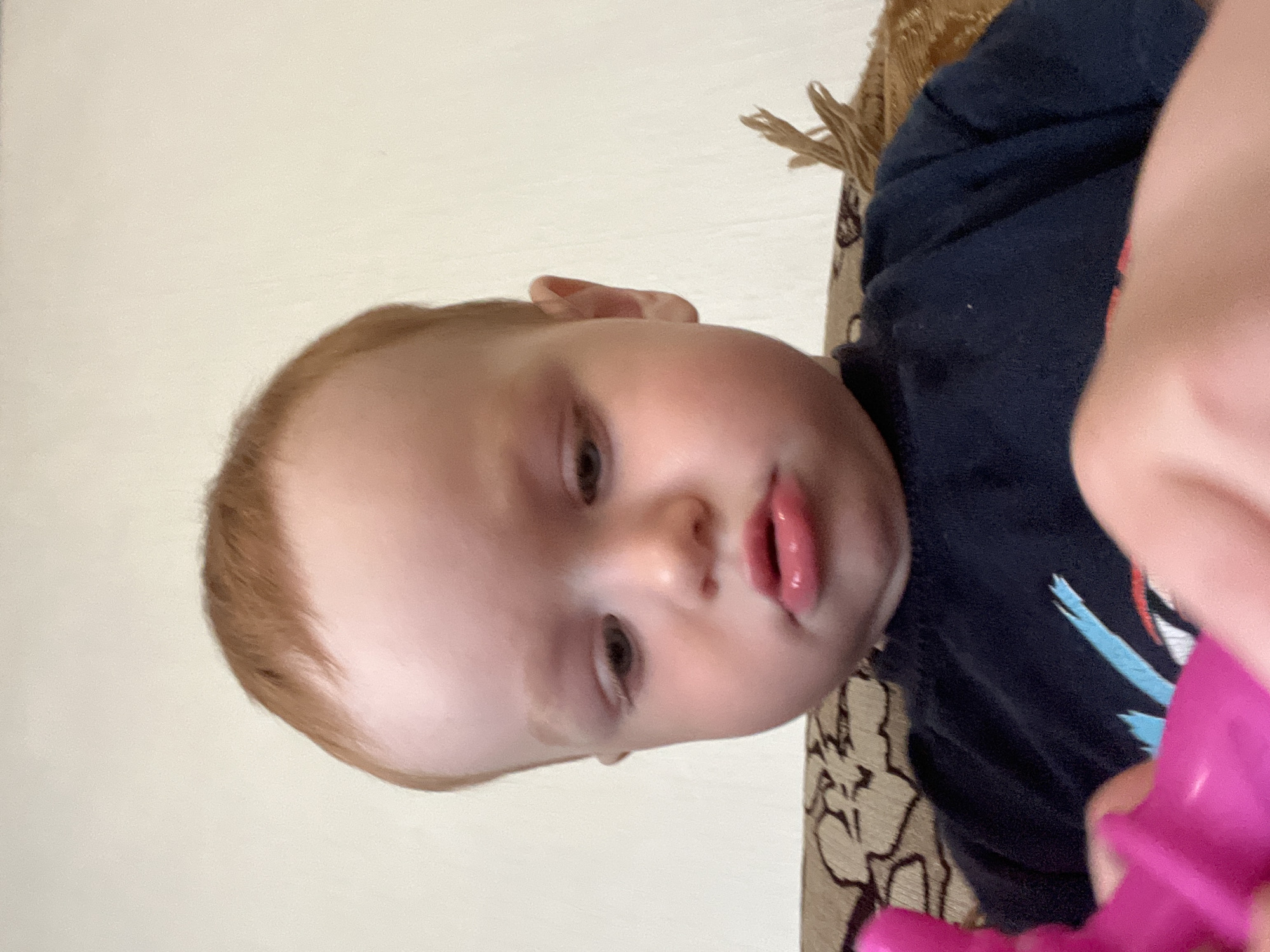 The image displays a young child, appearing to be a toddler, captured in a close-up, slightly downward-angled shot. The scene is located in Сосново, Russia.
The child has fair skin, reddish-brown hair, and a rounded face with full, rosy lips. The lower lip is noticeably extended in a pout, appearing glossy. The child's light-colored eyes are directed downwards and slightly to the viewer's left, indicating focus on an object below. The child wears a dark navy or black long-sleeved t-shirt that features a partial light blue and red graphic design on the chest.
Both of the child's hands are visible in the lower foreground, gripping a bright pink plastic toy. The specific type or function of the toy is not fully discernible, but it has a rounded shape and what appears to be a handle.
The immediate background consists of an upholstered piece of furniture, likely a couch or armchair, covered in a light brown fabric with a darker brown patterned design. To the right of the child's head, a fringed decorative element, possibly part of a cushion or throw, is visible. Behind the furniture, the wall is a plain, light-toned color, appearing off-white or cream, indicating an indoor setting. The lighting is soft and evenly distributed.
