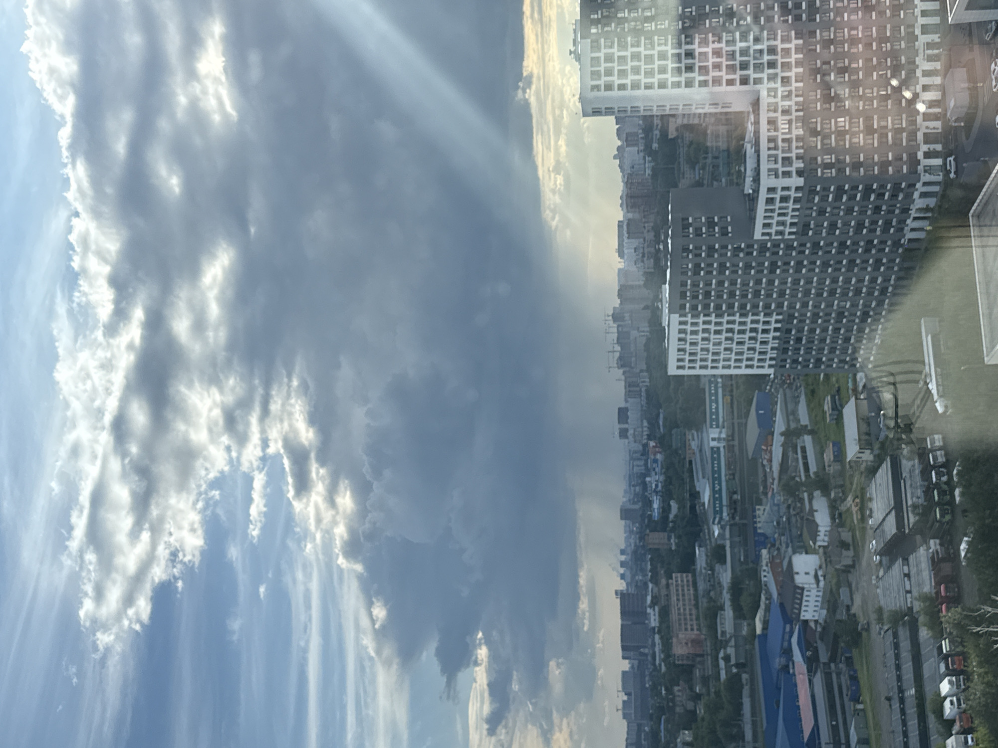 The image shows a wide shot of the city of Tomilino, Russia, taken from a high vantage point. The sky is dramatic, with a mix of bright sunlight and heavy, dark clouds. Sunbeams are piercing through the clouds, creating a dappled light effect over the cityscape. In the distance, a rain shower can be seen falling over a part of the city.
The urban landscape below consists of a variety of buildings, including tall, modern residential complexes with many windows, as well as lower-rise industrial or commercial buildings with flat roofs. There are also cranes visible, indicating ongoing construction. Green trees are interspersed throughout the city, especially in the foreground and around some of the buildings. In the lower part of the image, a parking lot or industrial area is visible with several trucks and white vehicles. The overall impression is of a busy, developing urban environment under a dynamic sky.