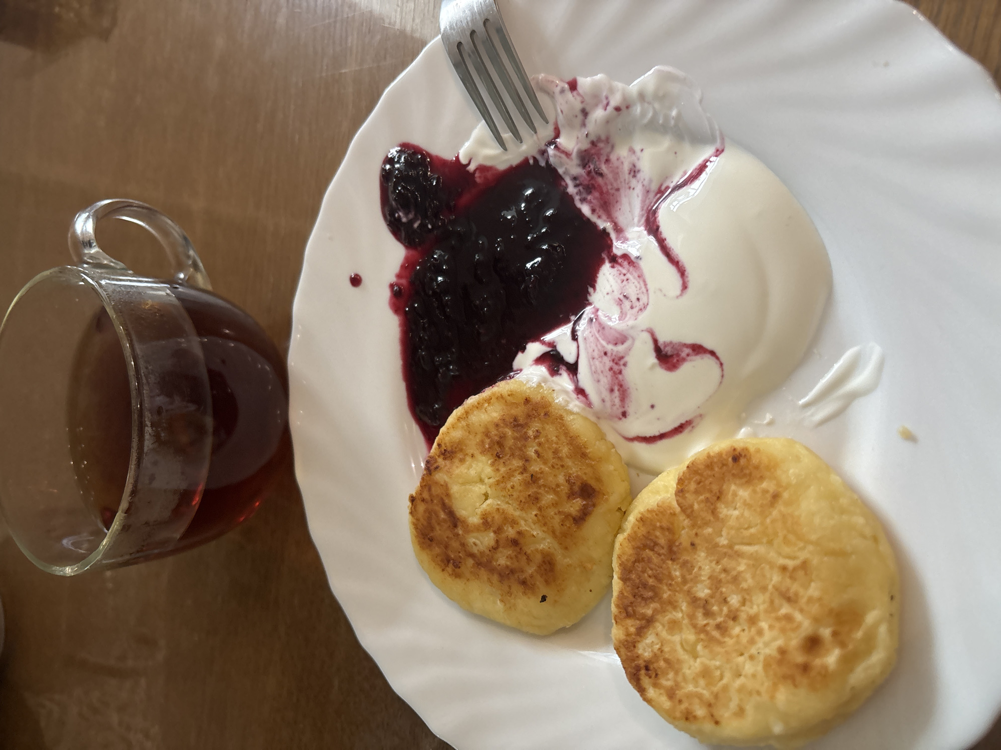 This scene depicts a breakfast or dessert meal served on a wooden table, located in the city of Chaikovsky, Russia. A white ceramic plate with a fluted rim occupies the lower and central portion of the frame. On the plate are two round, golden-brown pan-fried items, consistent with syrniki or cottage cheese pancakes, positioned on the left side. To the right of these items, a dollop of white cream, possibly sour cream or yogurt, is present, partially mixed with a dark purple, viscous berry jam or compote. A silver metal fork with four tines is positioned on the right side of the plate, partially submerged in the cream and jam mixture, suggesting recent use or preparation for consumption. In the upper background, slightly out of focus, is a clear glass mug with a handle, containing a dark reddish-brown liquid, likely tea. The wooden table surface beneath the plate and mug is a medium brown color, displaying subtle wood grain and a slight reflective sheen.