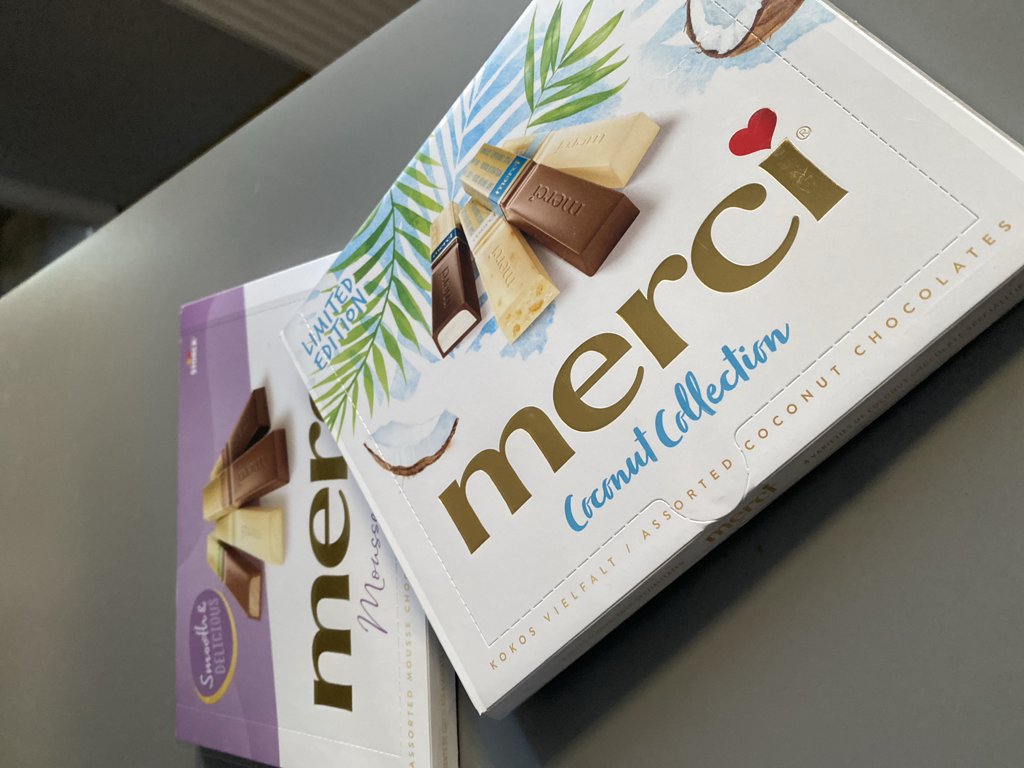 The image shows two boxes of Merci chocolates on a gray surface, possibly a table. The box in the foreground is white with "merci Coconut Collection" written in gold and blue. It also features an illustration of palm leaves and a coconut. The box in the background is purple and has "merci Mousse" written in gold. The chocolates themselves are not the primary focus but are visible on both boxes, hinting at the flavors within. The scene appears to be indoors, with soft lighting. There is no indication of the location being in Wrocław, Poland, or any other specific city.