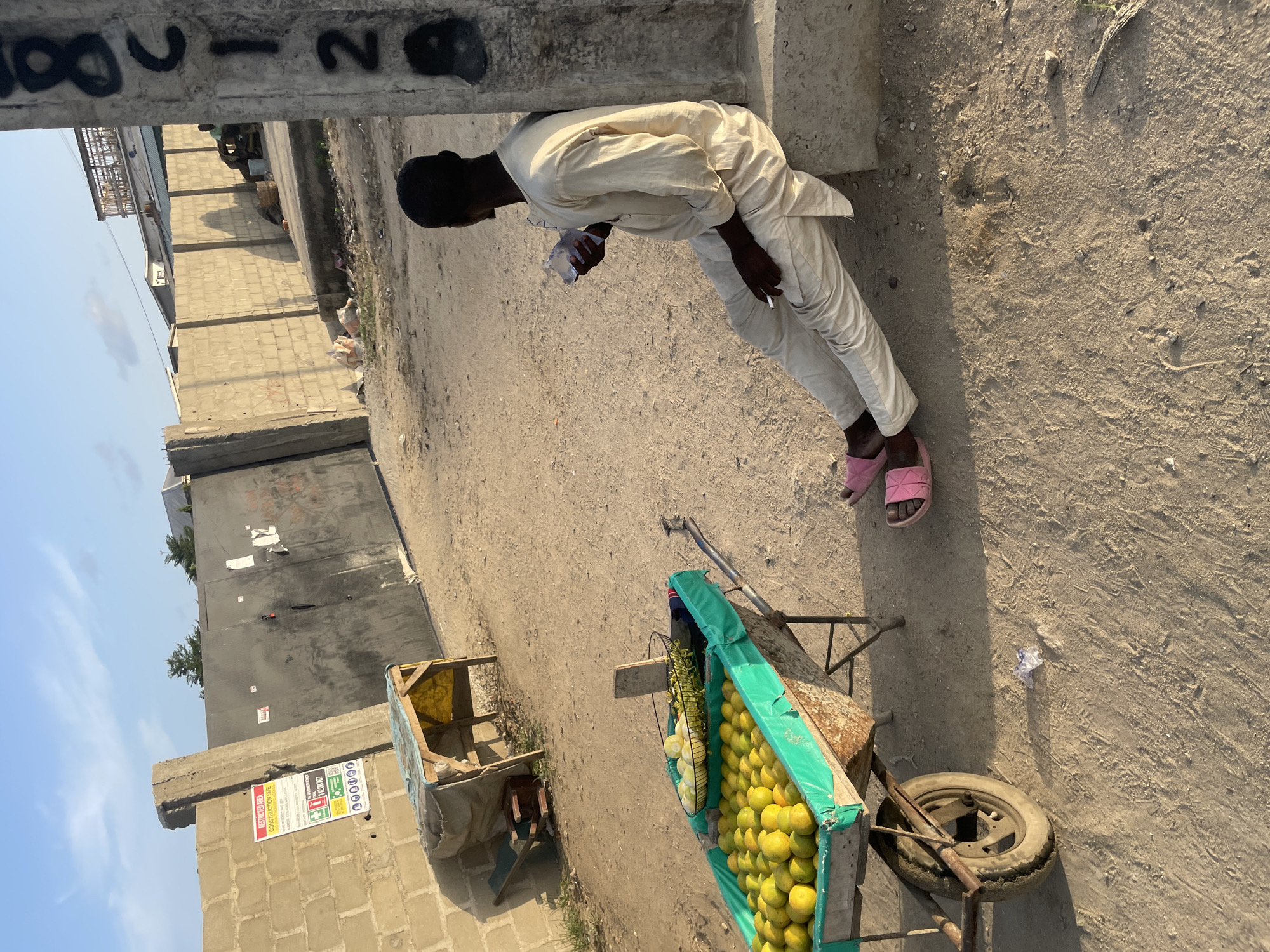 A man in a white outfit and pink sandals leans against a concrete structure on a dirt road. He holds a small plastic bag in one hand and appears to have a cigarette in the other. A cart filled with yellow fruit sits nearby, suggesting a fruit stand. Another cart is situated further down the road. The background shows buildings under construction and a clear sky. The location is Ado, Nigeria.