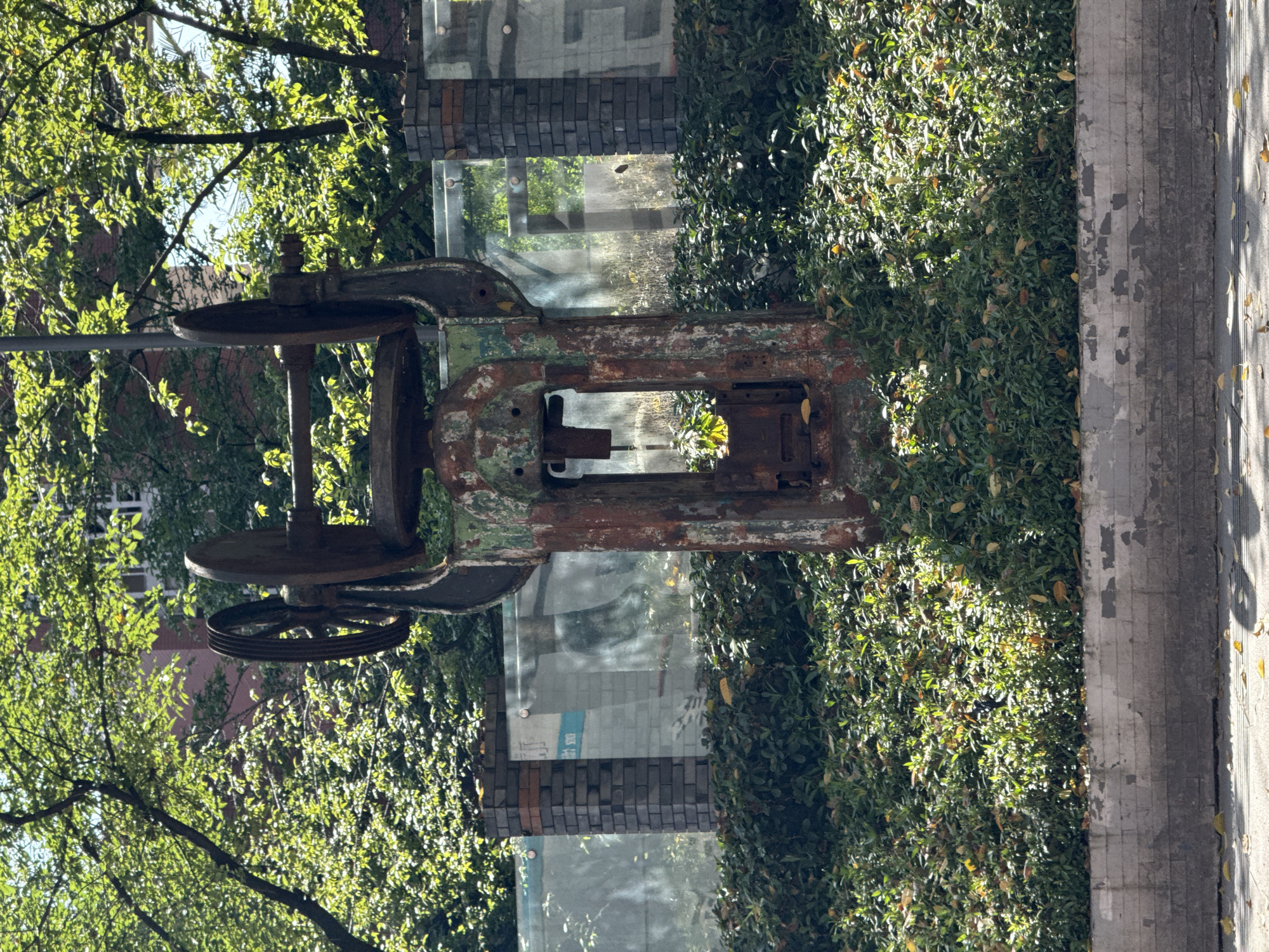The image shows a large, rusting piece of machinery in Jiulongpo District, China. The machinery consists of a large square frame with a rectangular opening in the center and two large wheels on one side. The machinery is positioned next to a sidewalk and a dense green hedge. A glass-enclosed structure with a brick base is visible behind the machinery. Trees with green leaves are present in the background and to the left of the machinery. The lighting is bright, with sunlight filtering through the trees.