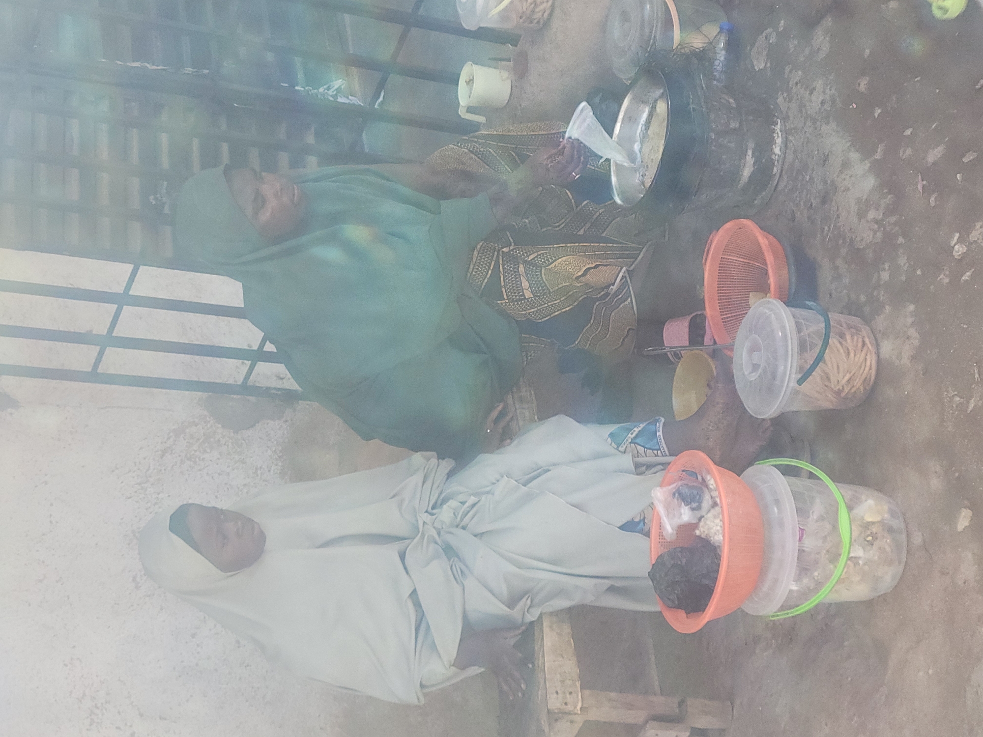 The image shows two women sitting on the ground in what appears to be an outdoor setting, likely in Jos, Nigeria, based on the clothing and general environment. One woman is wearing a light-colored hijab and robe, while the other is wearing a green head covering and a patterned robe. They are surrounded by various buckets and containers, some filled with food items or other materials. One woman is holding something in her hand, possibly a strainer or cloth, over a metal bowl. The ground is bare and somewhat dusty. The scene has a somewhat hazy or bright lighting. A metal railing is visible behind the women.