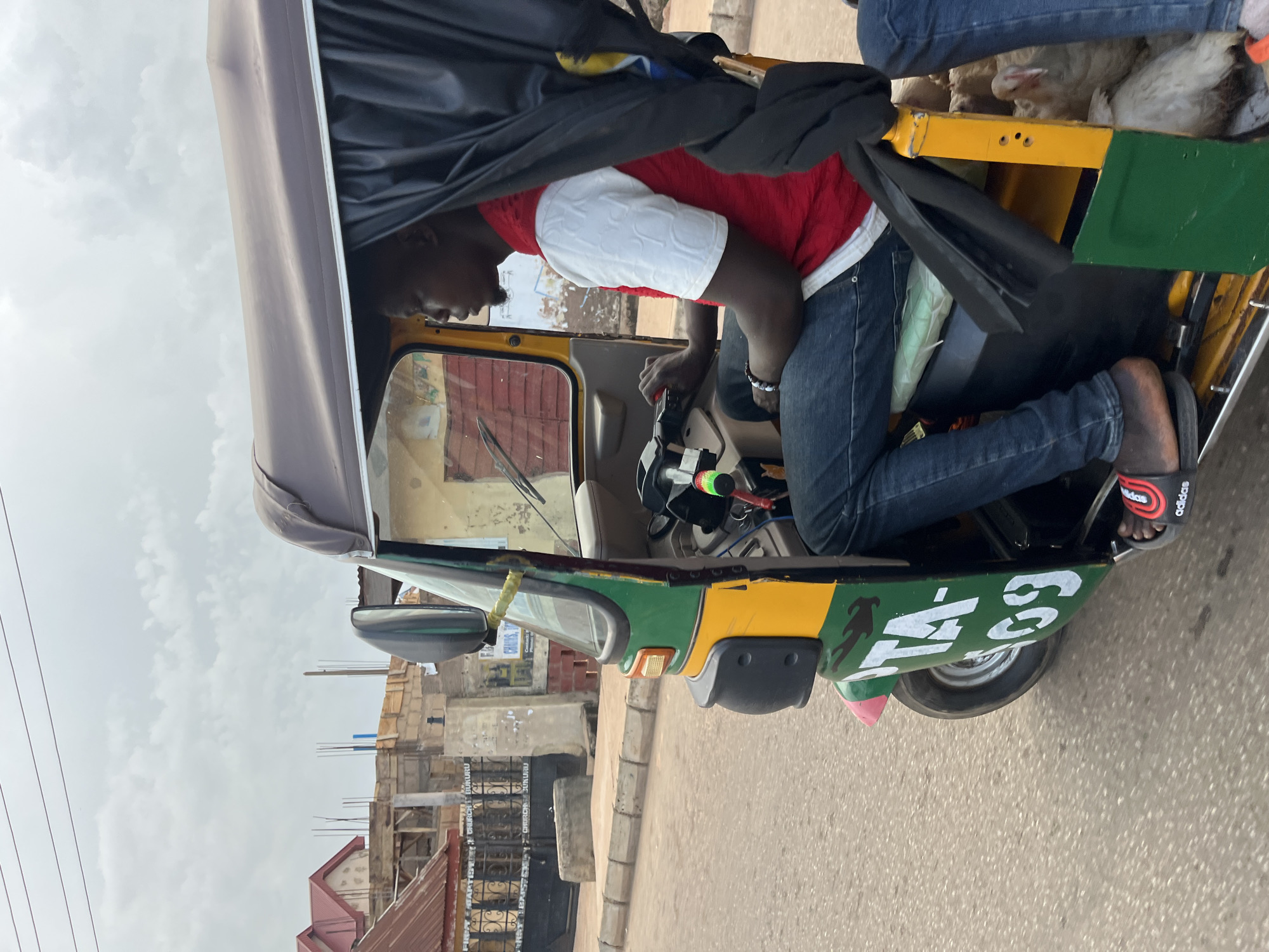 A man drives a yellow and green auto-rickshaw, also known as a "Keke," in Bukuru, Nigeria. The driver, wearing a white shirt with a red vest over it and blue jeans, is looking forward. The auto-rickshaw has black curtains on the sides and a brown roof. A white chicken is visible in the back of the auto-rickshaw, along with another person wearing blue jeans. The auto-rickshaw's side displays white lettering that reads "KTA-109". In the background, buildings and a gate with the words "FIRST BAPTIST CHURCH BUKURU" are visible. The sky is overcast with clouds.