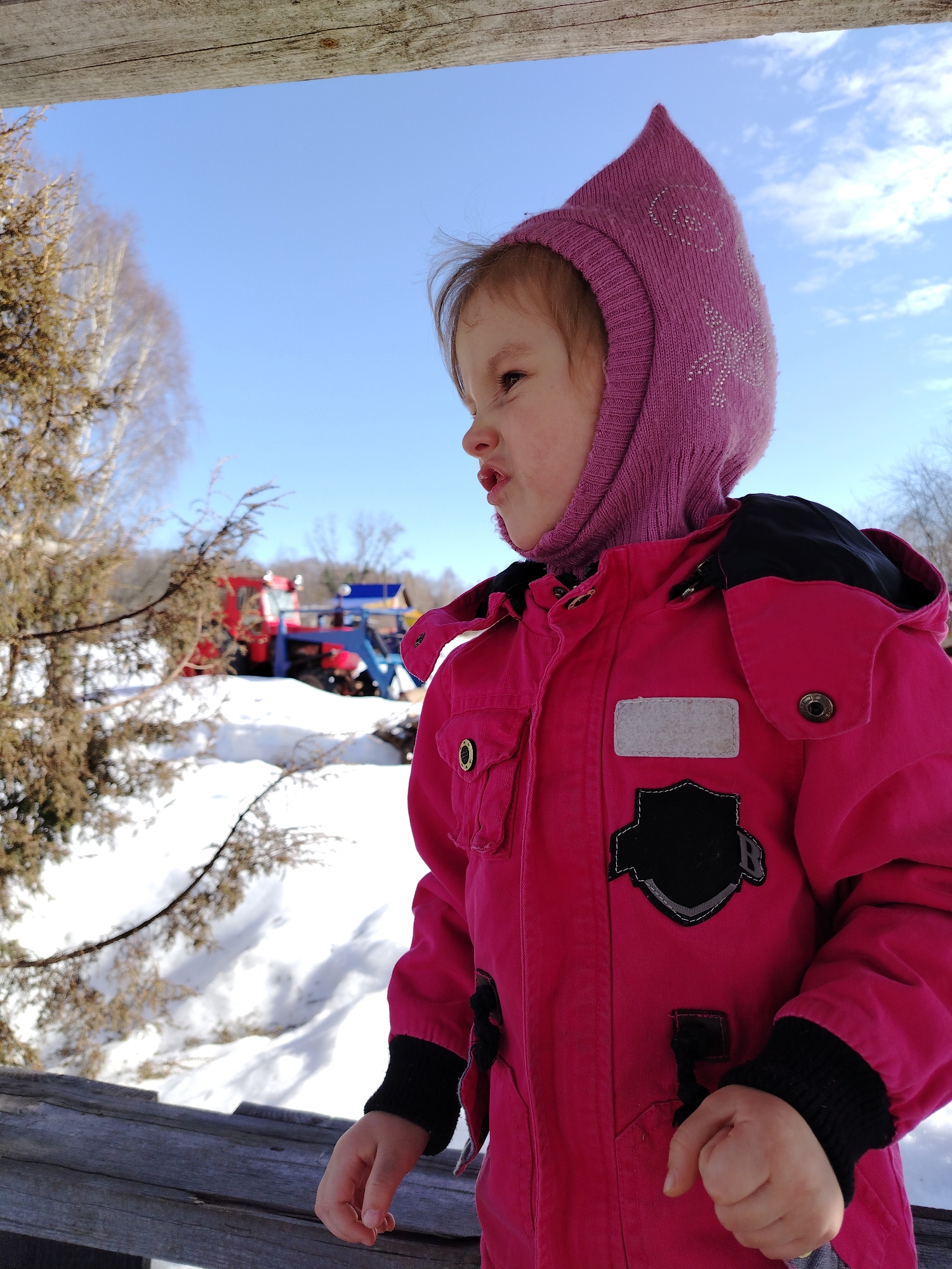The image captures a young child outdoors in Nizhnyaya Gar', Russia, under a clear blue sky with scattered white clouds. The child, seen from the mid-torso up, is positioned on the right side of the frame, facing left with pursed lips and a slightly upward-turned nose. The child is dressed in a bright pink winter jacket featuring black accents on the shoulders, black ribbed cuffs, a rectangular grey patch, and a black shield-shaped patch with white stitching and an 'R' on the chest. A pink knit hood, adorned with silver star-like embellishments, covers the child's head. The child's hands rest on a weathered wooden plank or railing, which runs horizontally across the bottom and partially up the left side of the frame. In the mid-ground and background, snow covers the terrain. Evergreen trees with brownish foliage and bare deciduous trees are visible. To the left of the child, a red vehicle, likely a tractor, equipped with a blue front-mounted attachment (possibly a snowplow or loader), is visible in the distance.