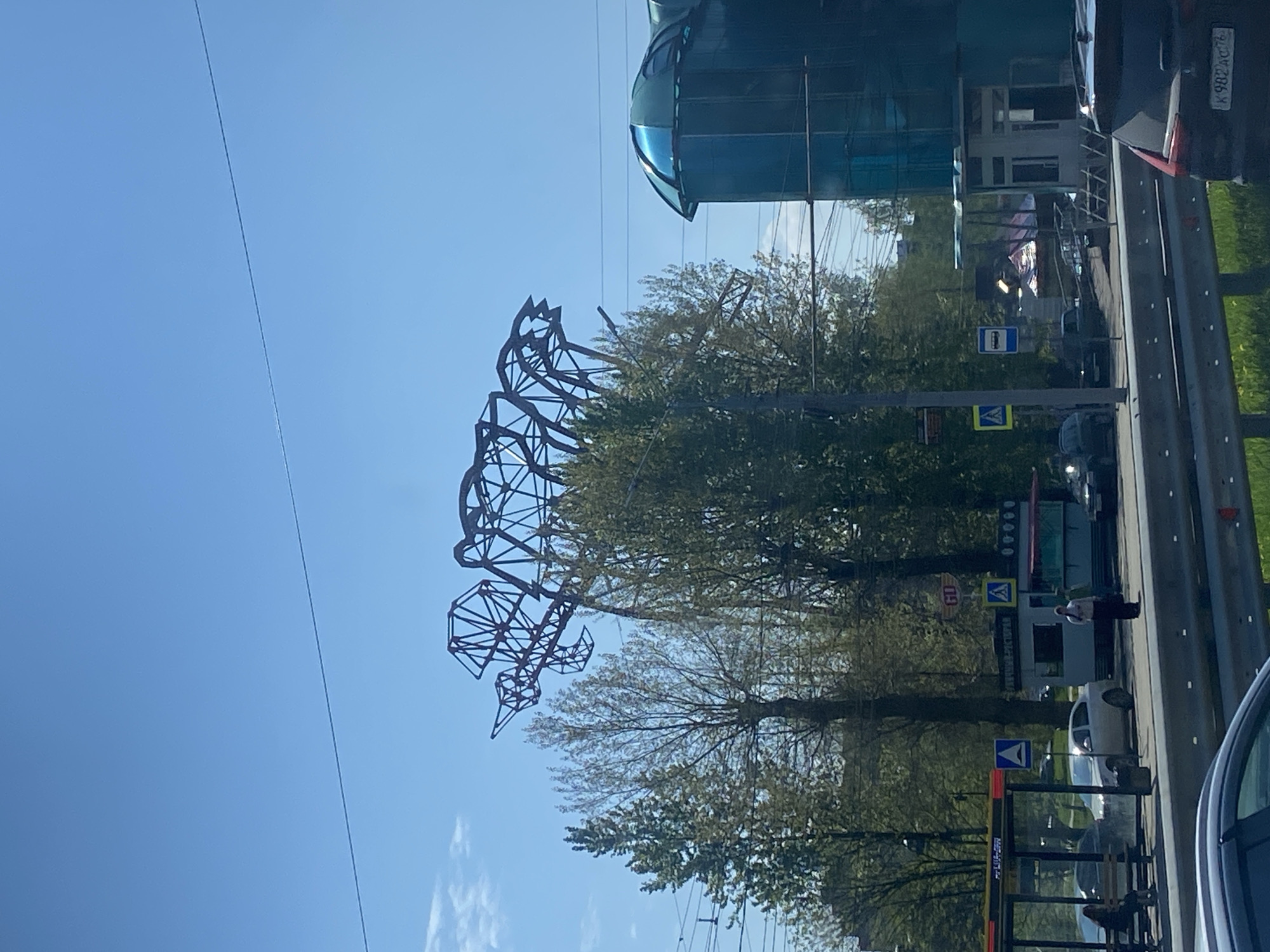 The image depicts a street scene in Rusanovo, Russia. On the left, a large metal sculpture of a bird is visible against a clear blue sky. Trees partially obscure the sculpture. To the right, a road with several vehicles, including a dark car with a license plate "K 982 AC 78", a white car, and a bus stop with pedestrians. The bus stop has signs, and a blue, modern building with a glass dome is in the background. The overall impression is that of a sunny day in an urban environment.