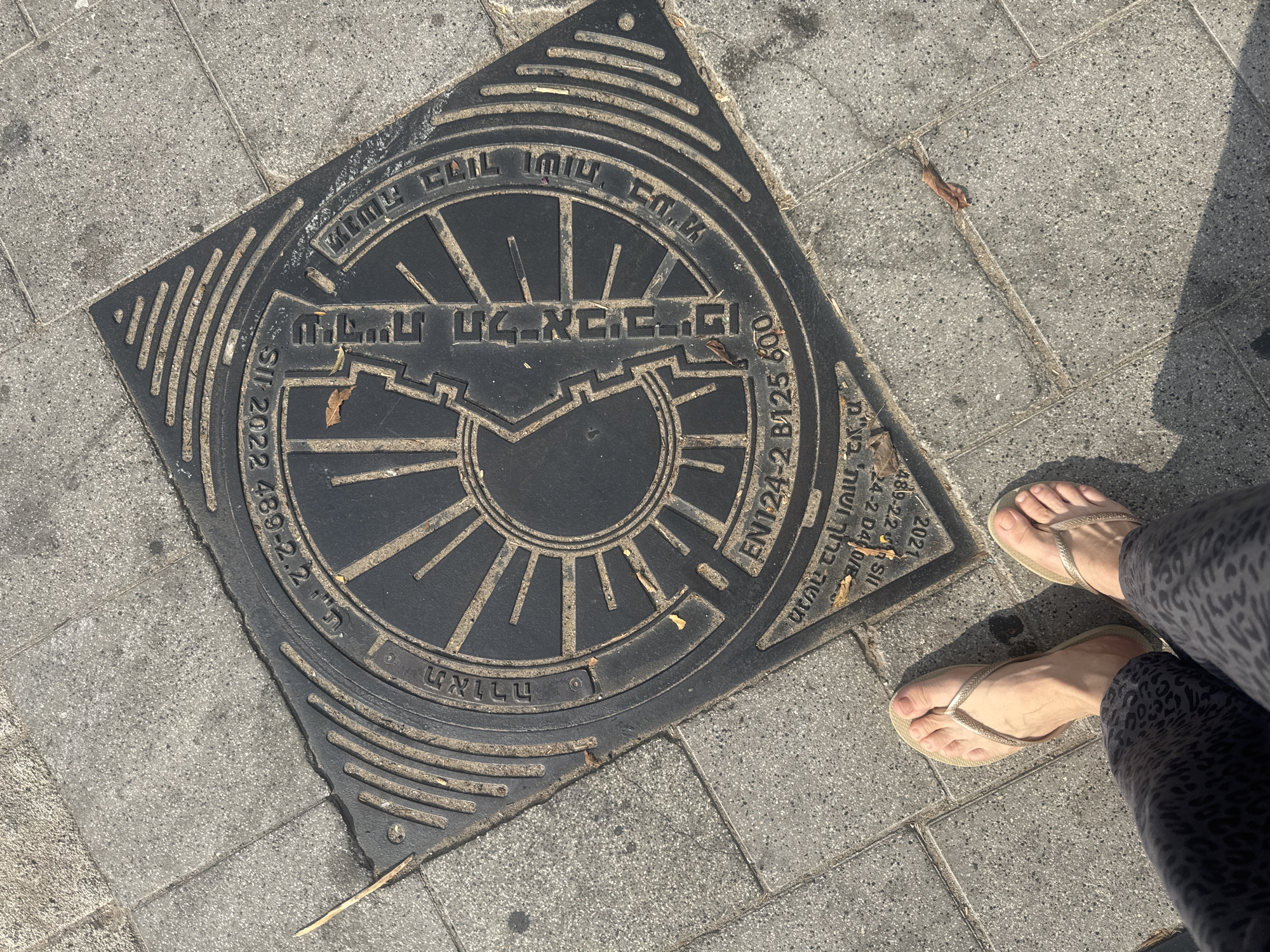 The image captures a view from a first-person perspective, looking down at a square manhole cover on a paved sidewalk in Tel Aviv, Israel. The manhole cover is made of black metal with raised lettering and a circular design featuring radiating lines. Text in Hebrew and English is visible on the cover, including "SIT 2022", "EN124-2 B125 600", and what appears to be a manufacturer's mark or standard specification. The sidewalk is composed of grey concrete tiles. Two feet wearing gold flip-flops are visible in the lower part of the frame, one foot wearing patterned leggings. The overall lighting suggests it is daytime with sunlight casting shadows.