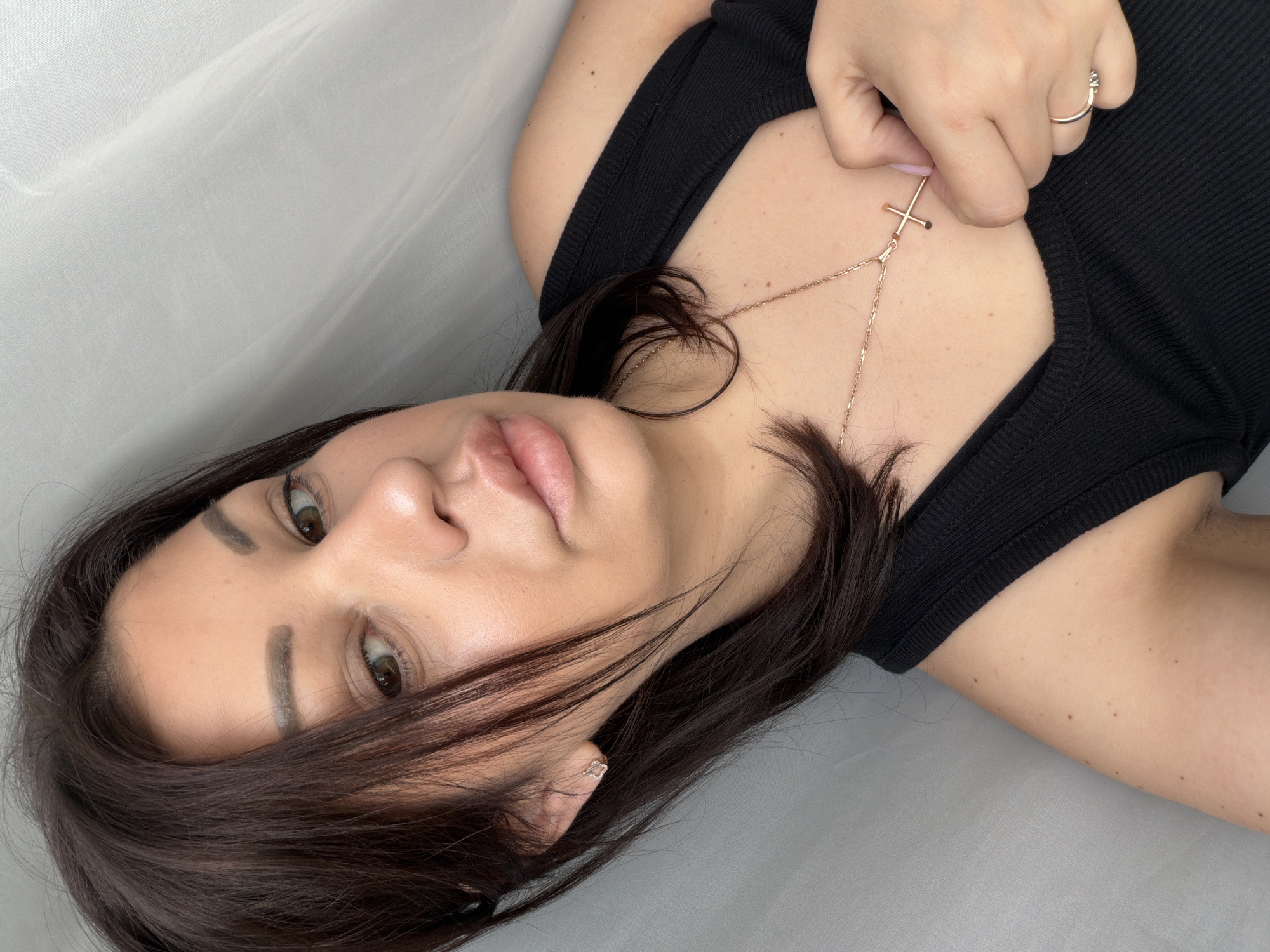 The image is a close-up selfie of a woman with dark brown hair and light brown eyes. She is wearing a black tank top and a gold cross necklace. Her left hand is resting on her chest, near the necklace. She is also wearing a small gold earring in her left ear and a gold ring on her right ring finger. The background appears to be a white, sheer curtain. There is no indication of the city of Sochi, Russia, in the image.