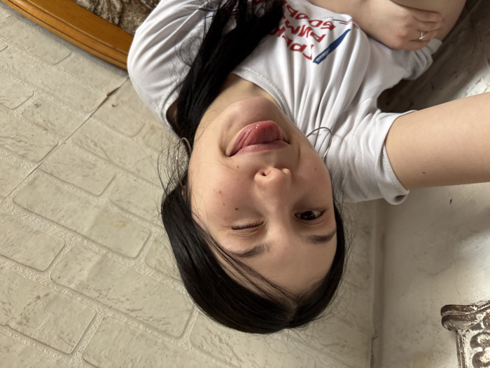 A female individual is captured in an indoor setting. She has dark hair, fair skin with visible freckles/moles on her left cheek, and is looking directly at the camera. Her right eye is closed in a wink, and her tongue is slightly extended from her mouth. She is wearing a light-colored, short-sleeved t-shirt with partially visible Cyrillic text in red and blue, including "ГРУППА КРОВИ". Silver-colored hoop earrings are visible on both ears, and a silver-colored ring is present on the fourth finger of her left hand. Her left arm is raised and bent, partially obscuring her midsection. The background to the right consists of a light-colored wall featuring a faux brick pattern. To the upper left, a section of a plain, lighter-colored wall is visible, with a dark, ornate, possibly metallic or carved wooden object affixed to it. A wooden furniture element, possibly a chair back or headboard, is partially visible in the lower right background. The specific location within the city of Фоки, Russia, is not visually determinable from the image.