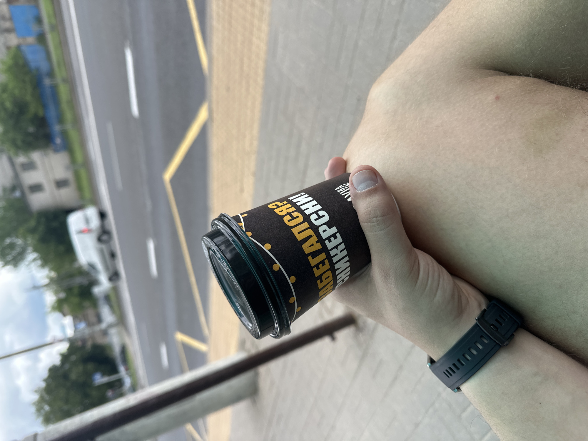 The image captures a close-up of a person's right arm and hand holding a black cup, set against a blurred urban background. A black digital or sports watch is visible on the person's wrist. The black cup features yellow text and small yellow dots, with the visible Cyrillic inscription reading "АБЕДАЙСЯ" and "СНИКЕРСНИ", which likely relates to a Snickers-flavored product or a playful slogan.
The background, significantly out of focus, reveals a multi-lane road with white lane markings and a solid yellow line along what appears to be a curb or sidewalk. A white van is partially visible and blurred on the road. Distant buildings and green trees are also indistinctly seen under an overcast or partly cloudy daytime sky. In the foreground, a dark metal railing or barrier is present, below which is a section of pavement with yellow tactile indicators, often found in public spaces such as bus stops or pedestrian crossings. The scene suggests an outdoor urban or suburban setting, likely in Minsk, Belarus, where the person is stationary, possibly waiting or resting by the side of the road.