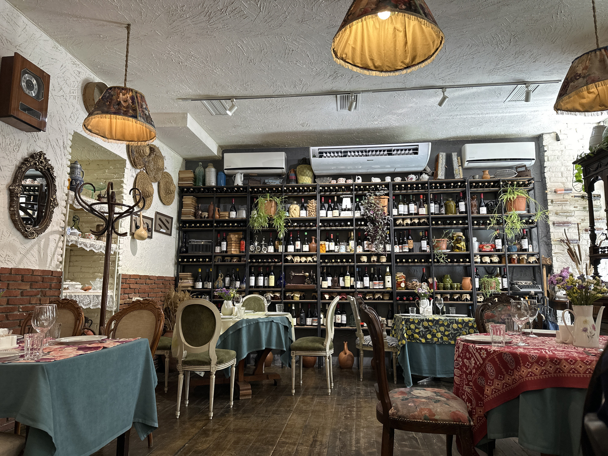 The interior of a restaurant in Tbilisi, Georgia, is depicted. The restaurant has a rustic style with exposed brick walls and wooden floors. Tables with chairs are arranged throughout the room. A large bookshelf filled with wine bottles and other decorative objects covers one wall. Three lamps hang from the ceiling. The lighting is warm and inviting. There are no people visible in the image.