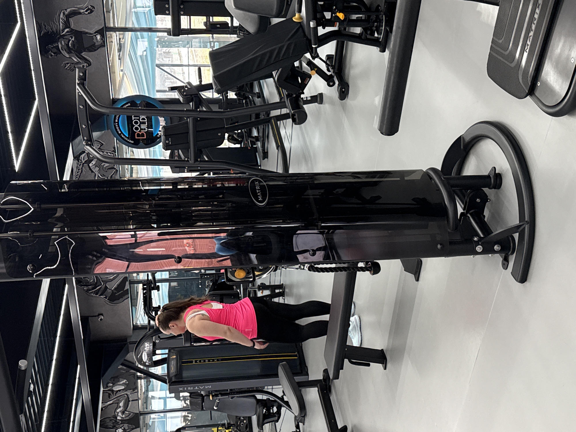 The image shows a fitness center with a woman exercising on a Matrix brand chest press machine. The woman is wearing a pink tank top and black leggings. Other exercise equipment is visible in the background. The location cannot be identified as Votkinsk, Russia, as the image lacks specific markers to confirm this location.