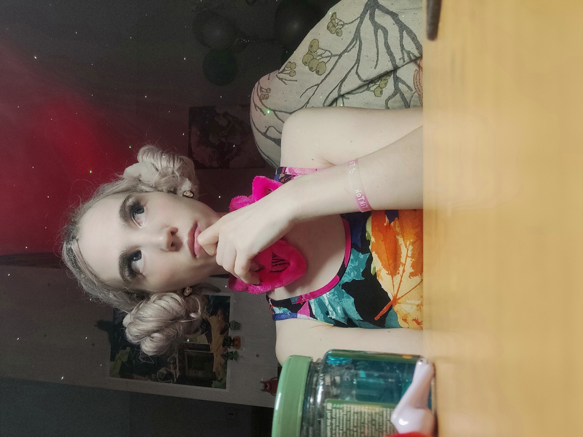 A young woman with pale skin and blonde hair styled in two buns poses for a photo. She is holding a pink, heart-shaped object to her lips. The woman wears a colorful tank top and a pink wristband labeled "TIKTOK." The background includes a wall with decorative elements, a patterned cushion, and a table covered with a jar and a small white object. The location is Chaikovsky, Russia.