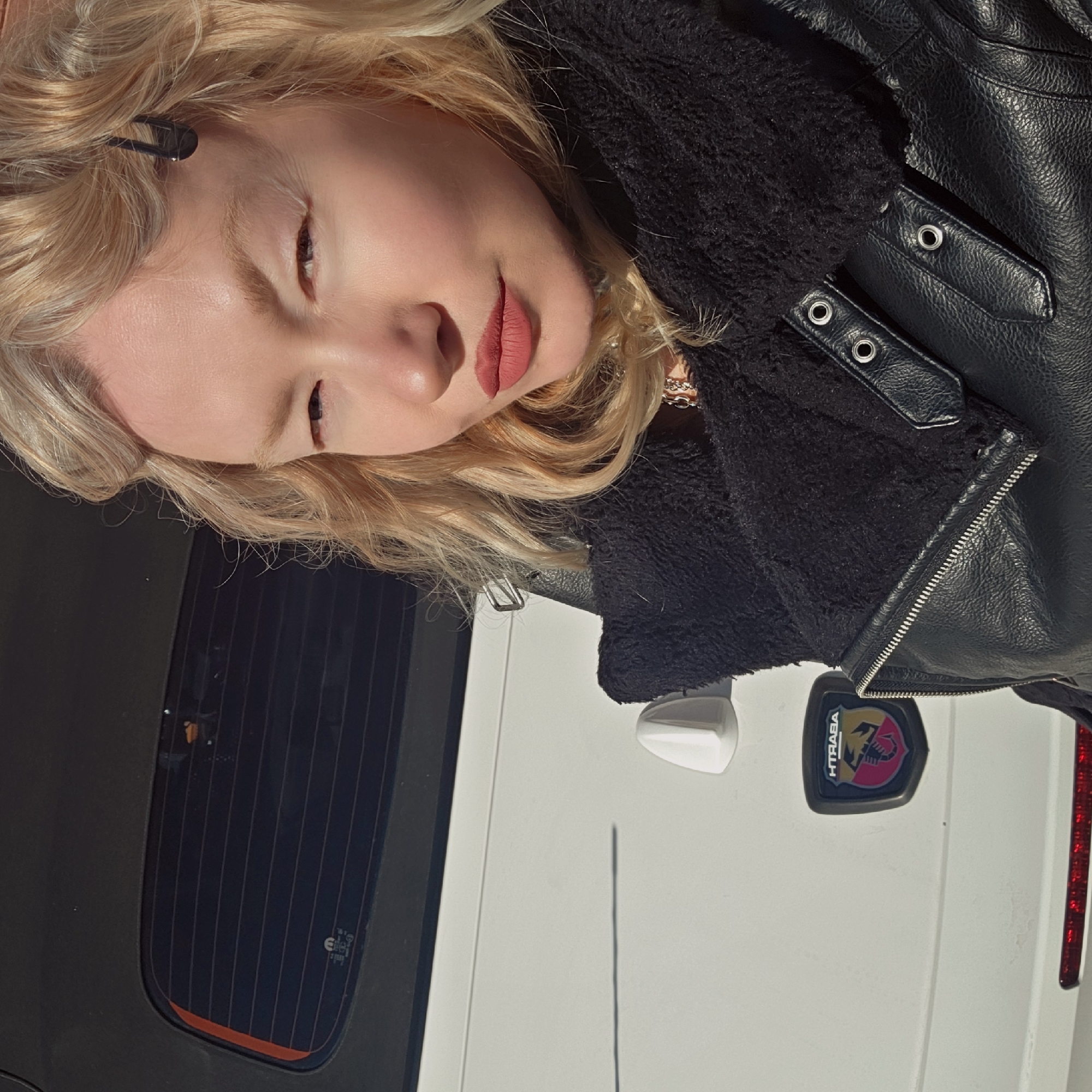 A young blonde woman wearing a black leather jacket with a furry collar is posing for a selfie. Her hair is wavy and styled with a dark bobby pin. She is wearing lipstick. Behind her, a white car with a black tinted rear window and an Abarth emblem is visible. The location is Saint Petersburg, Russia.