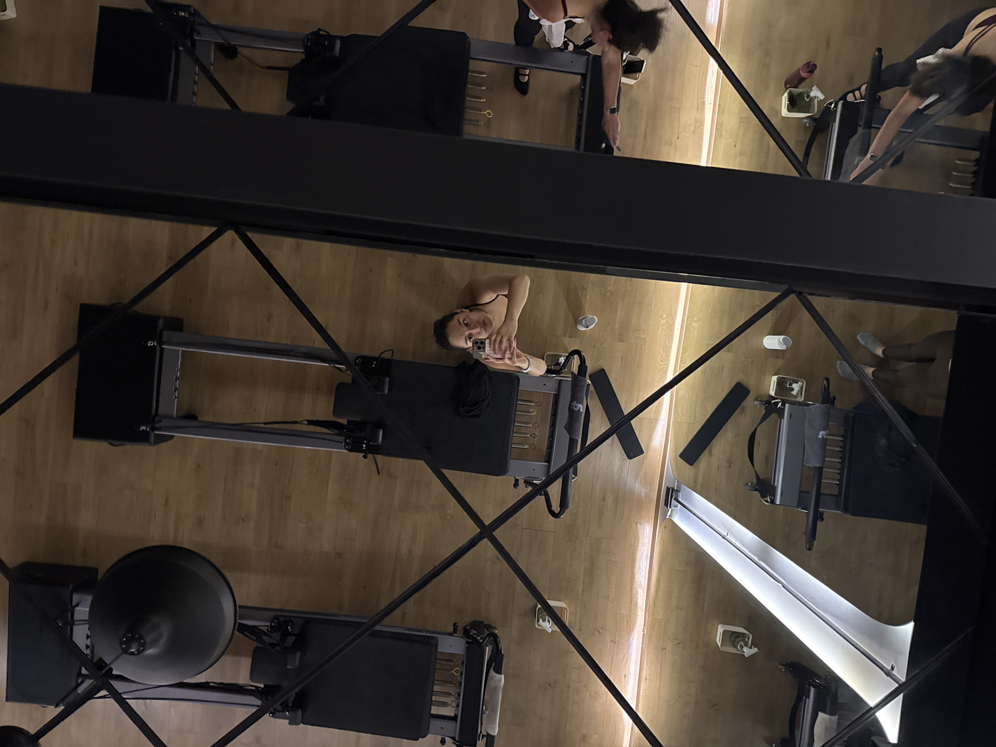 This is an overhead shot taken within a gym or fitness studio, specifically a Pilates studio. The flooring is light-colored hardwood. The shot includes multiple Pilates reformer machines. There are two women in the photo. One is operating a reformer machine, the second is facing upwards and appears to be taking a photo of herself. There are water bottles and personal items near the machines. The exact location within Puebla, Mexico cannot be determined from this image.
