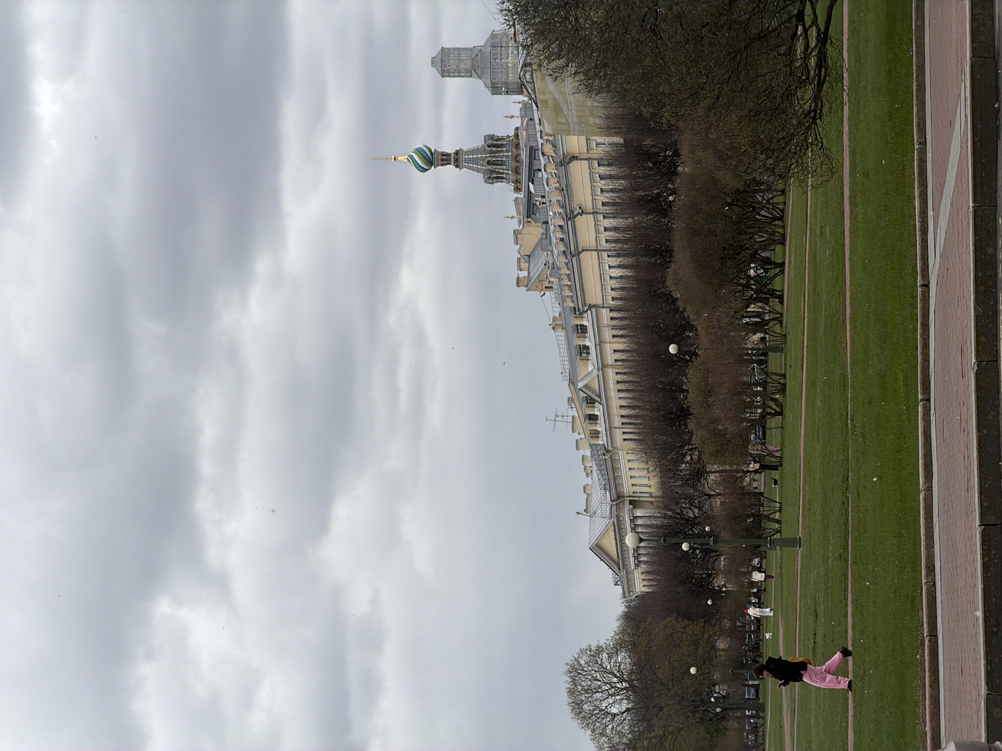 The image depicts an expansive outdoor scene in Saint Petersburg, Russia, likely a park or a large square, on an overcast day. The sky, which covers a significant portion of the upper frame, is filled with light grey, diffuse clouds, indicating a cloudy or muted daylight.
In the mid-ground, a prominent and ornate building stands out with a tall, slender spire capped by a distinctive onion dome adorned with blue, green, and gold stripes. This architectural feature strongly suggests the Church of the Savior on Spilled Blood. Adjacent to and behind this landmark are other classical-style buildings with multiple stories and numerous windows, showcasing a grand European architectural style.
In front of the buildings and extending into the foreground are vast, well-maintained green lawns, crisscrossed by faint lighter lines, possibly paths or divisions within the grass. Numerous deciduous trees with bare branches are scattered across the green space and line the area between the buildings and the lawns, indicating early spring or late autumn.
In the right foreground, a wide paved pathway made of reddish-brown and lighter-toned stone or brick, arranged in geometric patterns, runs along the edge of the green space. Several people are visible in the scene. In the immediate foreground, a person wearing a dark top, a dark jacket, and bright pink pants walks across the grass near the paved area. Further back on the lawns, several smaller figures can be seen, some standing and one appearing to wear white. Spherical white streetlights are also visible throughout the park area. The overall atmosphere is tranquil under the soft, diffused light of the cloudy sky. There is no visible text in the image.