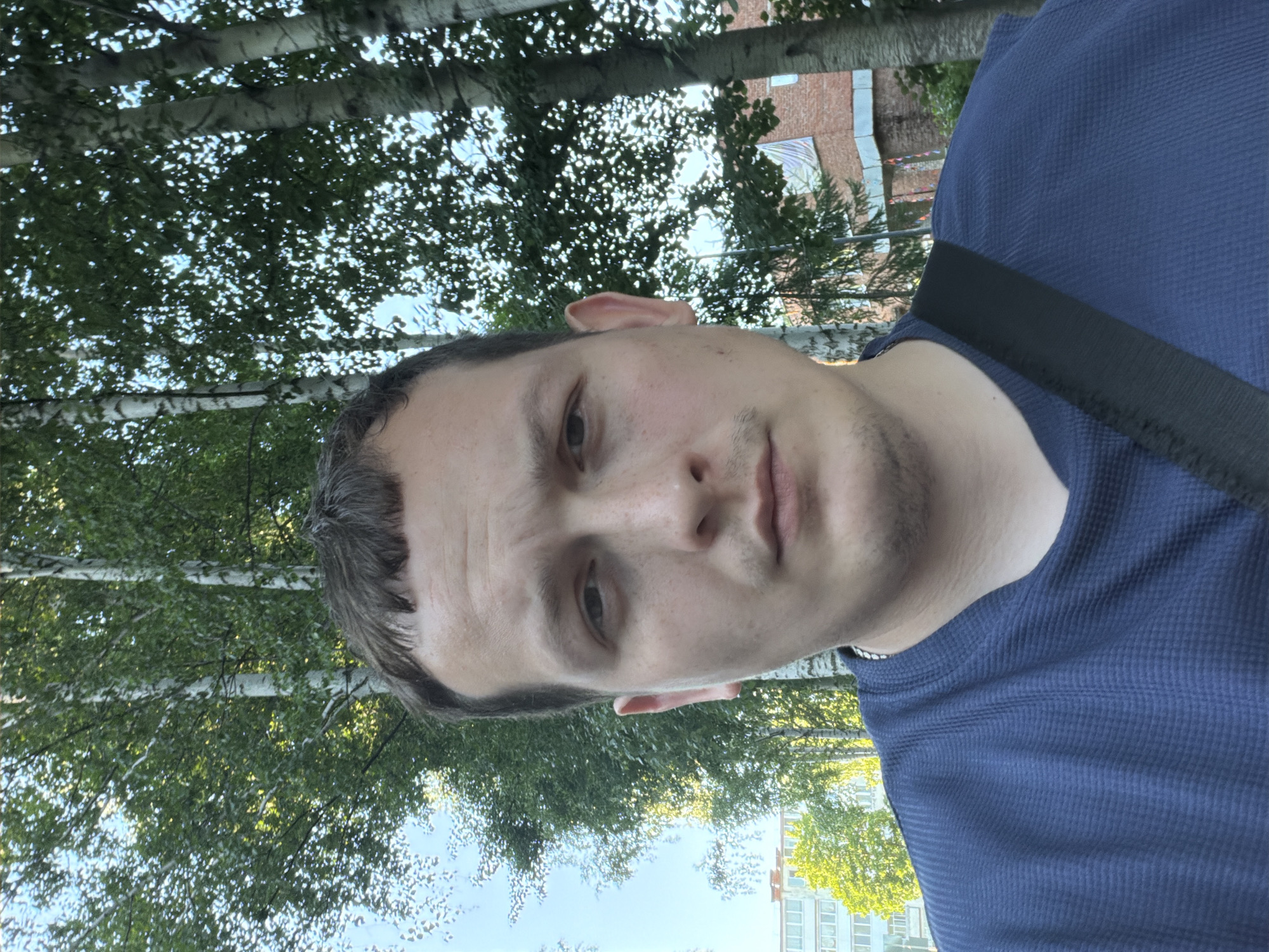 The image shows a close-up, selfie-style photograph of a person's face and upper body. The individual appears to be a male with short, dark hair, fair skin, and light-colored eyes. He is wearing a dark blue, textured t-shirt. A black strap is visible across his right shoulder. He is outdoors, with a background of trees, specifically birch trees, and foliage. In the distance, there are buildings, one of which appears to be a light-colored, multi-story structure, and another with brickwork. The sky is visible and appears clear. The lighting suggests it is daytime, with sunlight filtering through the leaves of the trees. The specific location in Noviy, Russia, is not discernible from the visual information alone.