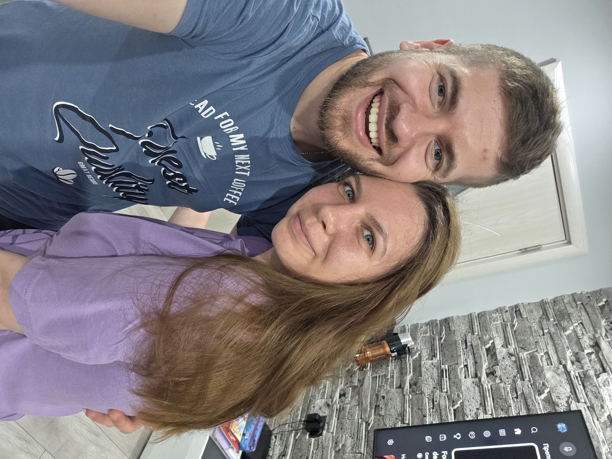 A man and a woman are posing for a selfie. The man is wearing a blue t-shirt with a coffee-related design. The woman is wearing a light purple t-shirt. They are indoors, with a wall featuring a textured, brick-like design behind them. A television is visible in the lower part of the frame, along with some objects on a stand near the TV. The location, Yagodovo, Bulgaria, is not discernable from the image.