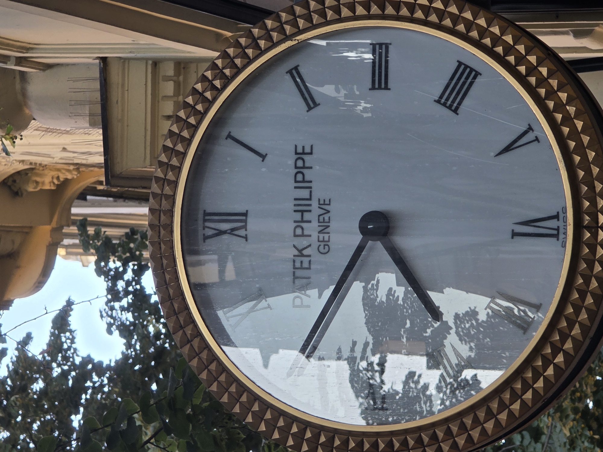A close-up shot of a Patek Philippe watch face is featured, framed by a decorative gold bezel adorned with pyramid-like studs. The watch has a light-colored face with Roman numerals marking the hours. The hour hand points to approximately VI, and the minute hand points to approximately IX. Reflections of trees and sky are visible on the glass surface of the watch face, creating a somewhat hazy effect. Behind the watch, architectural elements and foliage can be seen, suggesting an outdoor setting in a city.