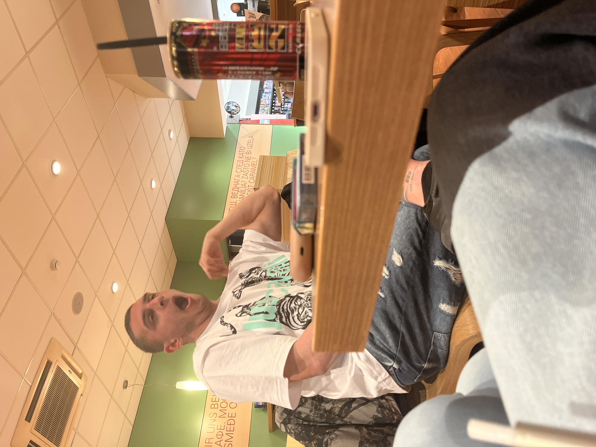 The image depicts an indoor scene within what appears to be a cafe or fast-food establishment in Burgas, Bulgaria. The main subject is a man with short dark hair, wearing a white t-shirt featuring a black and teal tiger graphic, and camouflage-patterned pants. He is seated at a wooden table, captured in a moment of strong emotion, looking upwards and to his right with his mouth wide open, suggesting surprise, excitement, or shouting. His left arm is bent, hand slightly clenched, indicating an animated gesture.
Another person is partially visible in the bottom right, seen from the waist down, wearing light blue jeans and dark clothing, also seated at the wooden table. On the table, a red energy drink can with a black straw is present, bearing the prominent text "K27" on its label, resembling a Red Bull can. A smartphone or similar device lies next to the can, partly obscured by the table structure.
The background shows a light-colored tiled ceiling with multiple recessed lights and a large air conditioning unit. A section of the wall is painted green. Visible text on the wall, in Cyrillic script, includes phrases like "РЕШ, ВЕДНАГА СЛЕД КАТО" (Decide, immediately after), "ANILIA? ZAŠTO NE BI UZELA" (Anilia? Why wouldn't she take), and "SIST CARAMEL?" (Is it caramel?), likely referring to menu items in Bulgarian or a similar Slavic language. Other partial text in Latin script, such as "IR UNS BE" and "AФЕ. MO," could suggest a multilingual setting. The lighting suggests it is daytime or a brightly lit interior.