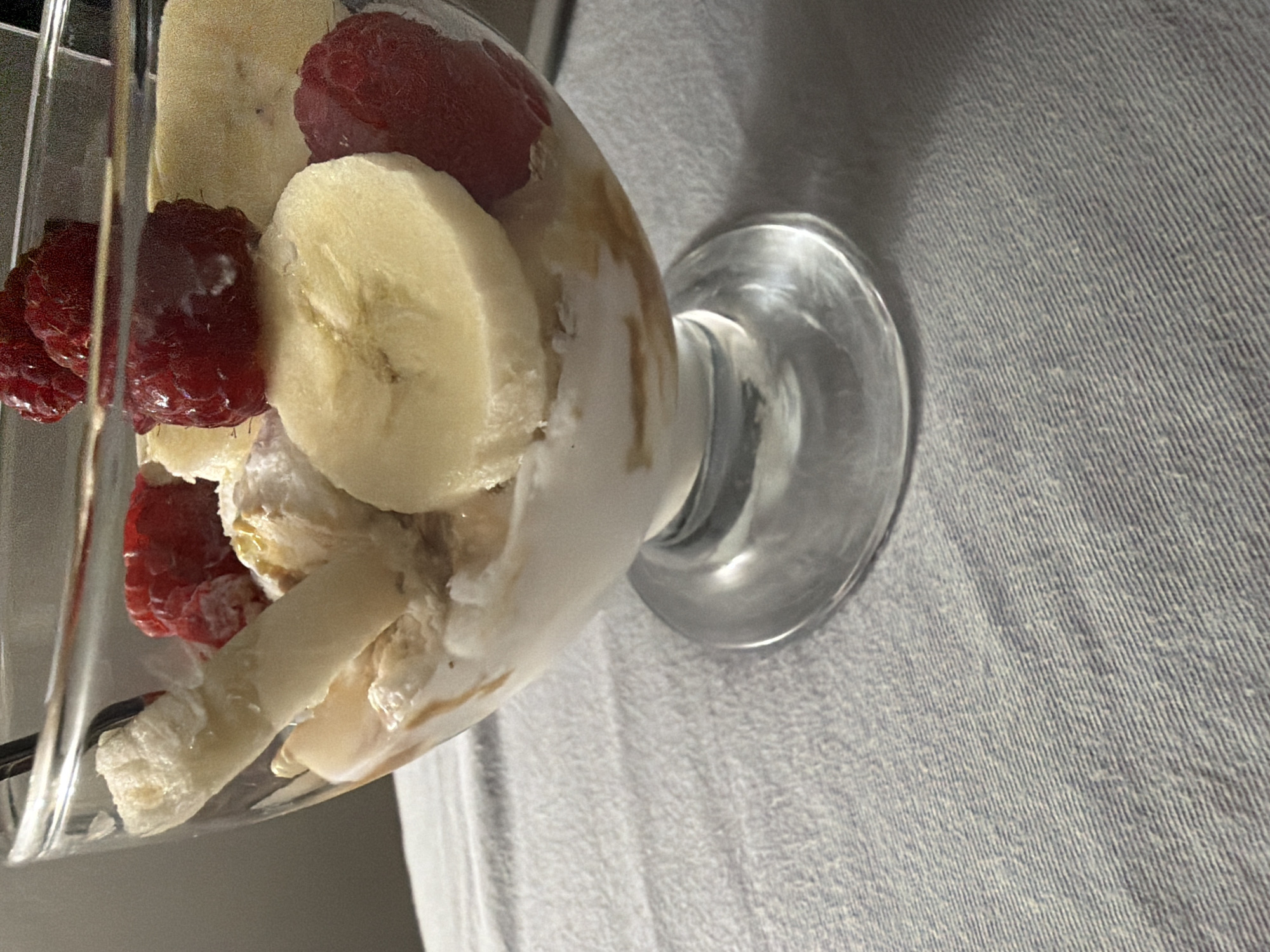 A close-up, eye-level shot shows a glass dessert bowl filled with ice cream, sliced bananas, and raspberries. The bowl is clear glass with a stemmed base and sits on a light blue, textured tablecloth. The ice cream appears to be vanilla, with streaks of caramel or syrup. Several slices of ripe banana are visible, along with bright red raspberries. A spoon is partially submerged in the dessert. The background is a plain, muted gray wall. There are no people in the image, and no identifiable landmarks or elements that would indicate its location in Pazardzhik, Bulgaria.