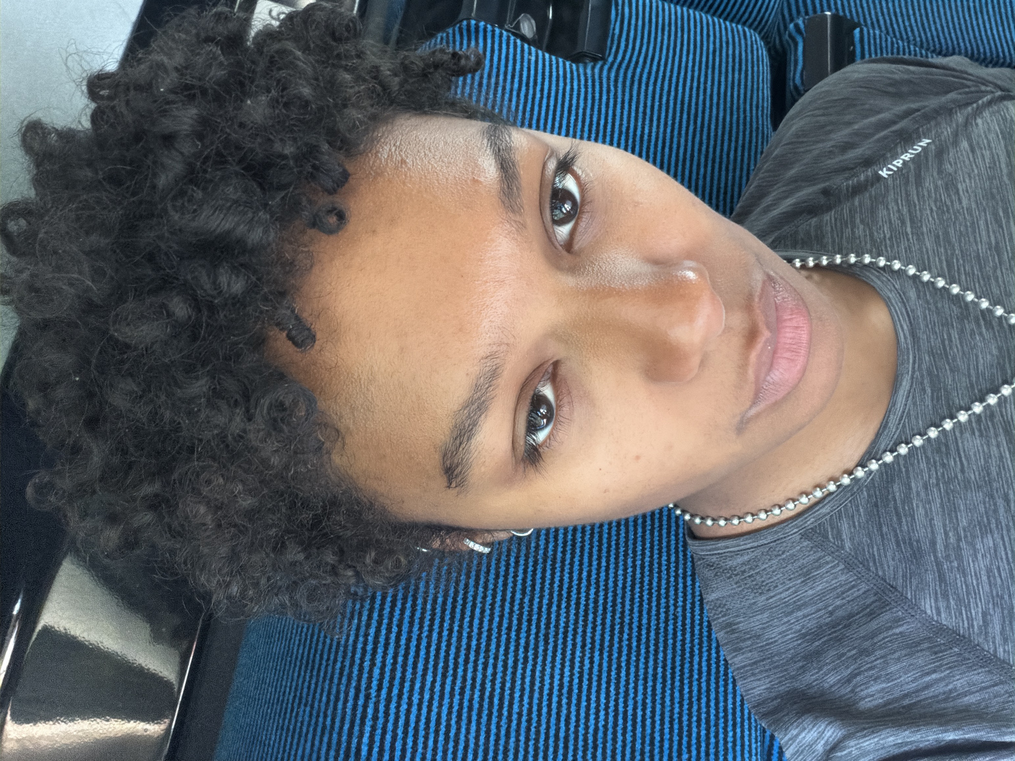 A selfie taken from a low angle, featuring a person with short, dark, curly hair, light brown skin, and dark eyes. The person is wearing a gray shirt with a "KIPRUN" logo and a silver beaded necklace. An earring is visible in the left ear. The background is blue with black stripes and black elements are to the left. The location is unknown and there is no information about Lormont, France.