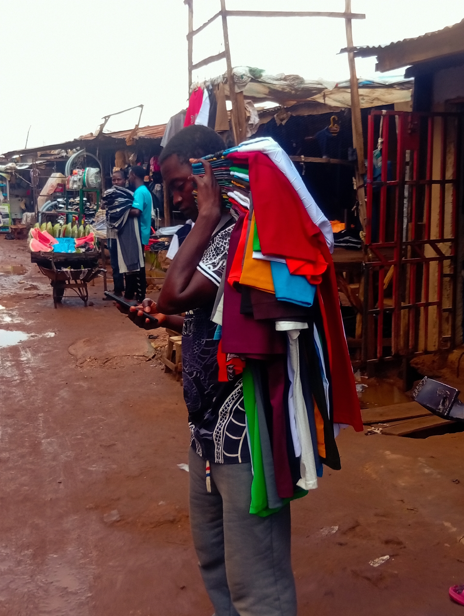 The image depicts a street market scene in Bukuru, Nigeria. The main subject is a person, likely a vendor, carrying a large bundle of colorful folded clothes on their left shoulder, which extends down their back. They are also holding a smartphone in their right hand, looking down at it. The vendor is wearing a dark, patterned t-shirt and grey trousers.
In the background, other market stalls are visible, constructed with corrugated metal roofing and wooden supports. One stall has a rack of red and white clothing hanging. Further down the path, another individual in a bright turquoise shirt stands near a cart piled with what appears to be fruit, possibly plantains and sliced watermelons. The ground is a reddish-brown dirt, wet and muddy in places, suggesting recent rain or standing water. The overall lighting suggests it might be daytime, possibly overcast. The atmosphere appears to be that of a busy, functional market. There is no visible text in the image.