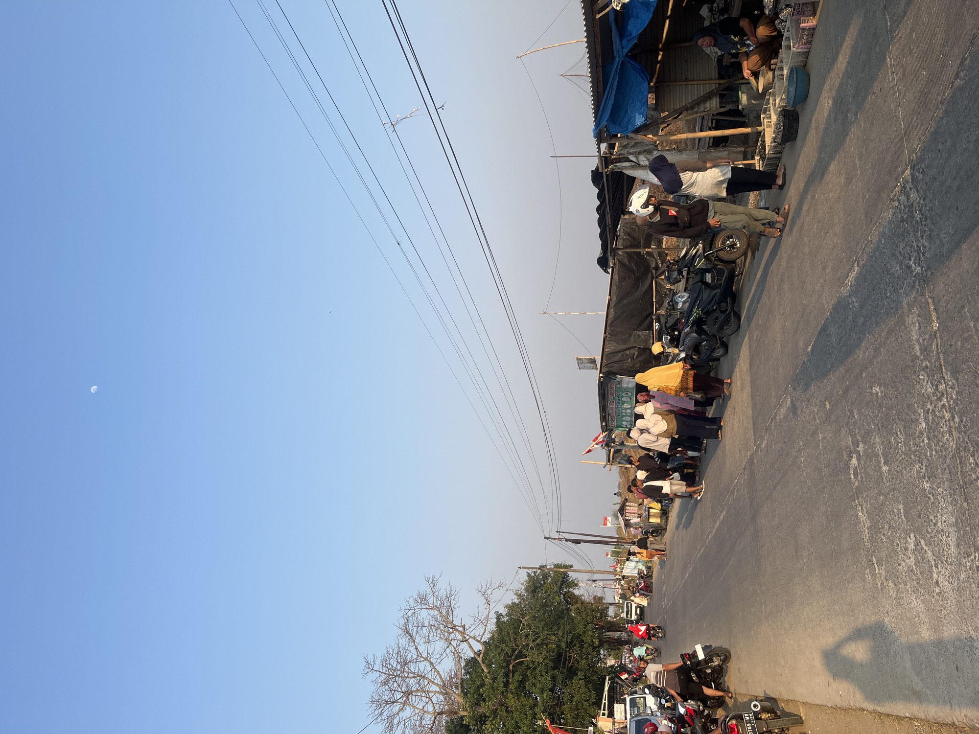 The image depicts a street scene in Kedungmalang, Indonesia, likely a market or roadside gathering. The clear blue sky features a faint crescent moon. A paved road runs through the center of the frame, lined with various stalls and parked motorcycles. On the left side of the road, there is a large tree with sparse foliage and several parked cars and motorcycles. A line of people, mostly women wearing hijabs of various colors, are walking or standing along the right side of the road, near the stalls. The stalls appear to be covered with tarps and corrugated metal, selling what seem to be food items, possibly seafood, displayed in crates. Several individuals are interacting with the vendors. The lighting suggests it is either early morning or late afternoon, with long shadows cast across the road. Indonesian flags are visible on some of the stalls.