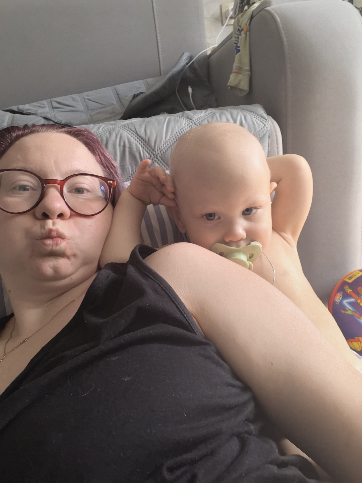 In an indoor domestic setting, likely a living room or bedroom in Chaikovsky, Russia, a woman with burgundy-dyed hair and red-rimmed glasses is taking a selfie while lying down. She is wearing a black tank top and has her lips puckered as if for a kiss. Her arm is around a baby who is also lying down. The baby has a pacifier in its mouth and is looking directly at the camera. The background consists of a sofa with a grey quilted blanket and a glimpse of a toy.