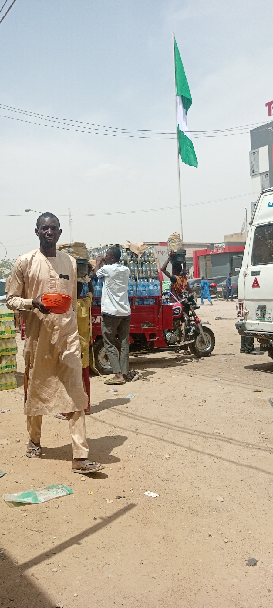 A street scene in Fagge A, Nigeria, features a man in a light brown tunic and pants holding an orange bowl. Behind him, other men load bottled water onto a red three-wheeled vehicle. A Nigerian flag is displayed. A white van and a building with "TAJBank ATM 24/7" sign can be seen in the background. The ground is dirt with some scattered litter.