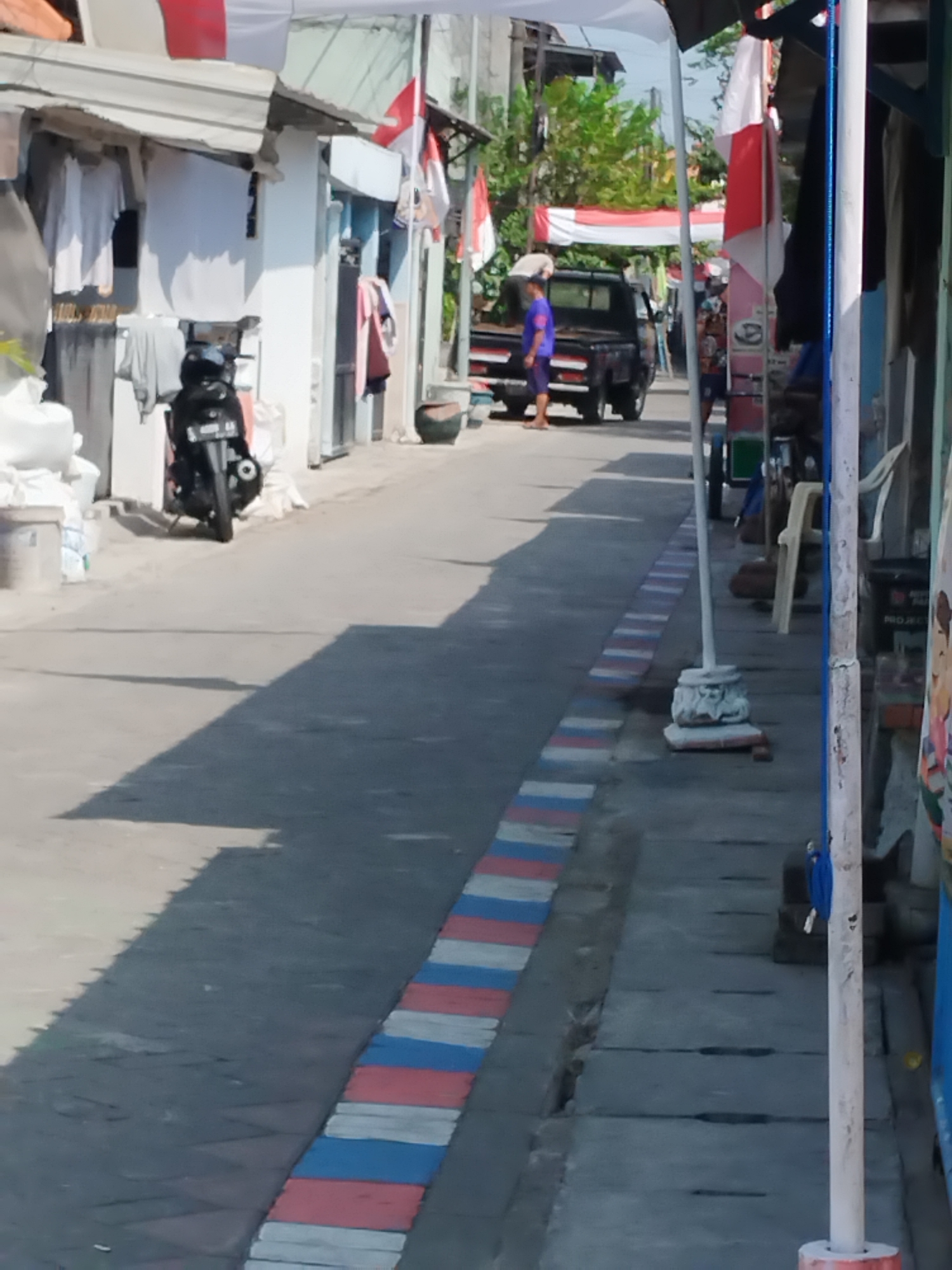 The image depicts a narrow street in Surabaya, Indonesia, characterized by residential buildings with overhanging awnings and laundry hanging out to dry. A black motorcycle is parked on the left side of the street. In the distance, a person in a blue shirt stands near a black pickup truck. Red and white flags, possibly indicating a national holiday, are displayed on some of the buildings. The street itself is paved and features a decorative border of red, white, and blue blocks. Shadows suggest a bright, sunny day.